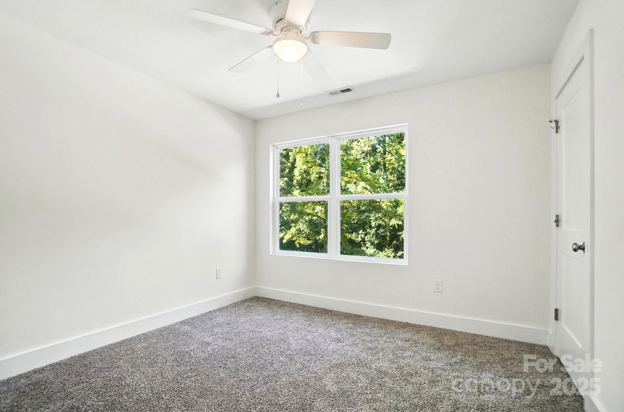 Empty bedroom with gray carpet, a ceiling fan, and a window overlooking leafy trees.