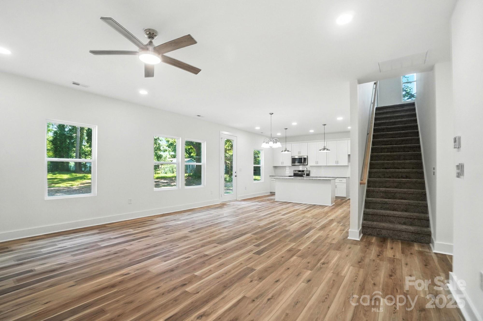 Open living area with wood-look floors, white kitchen island, large windows, and carpeted staircase.