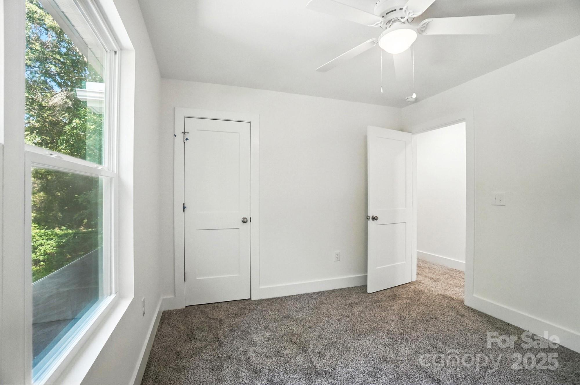 Empty carpeted bedroom with a large window overlooking trees and a white ceiling fan.