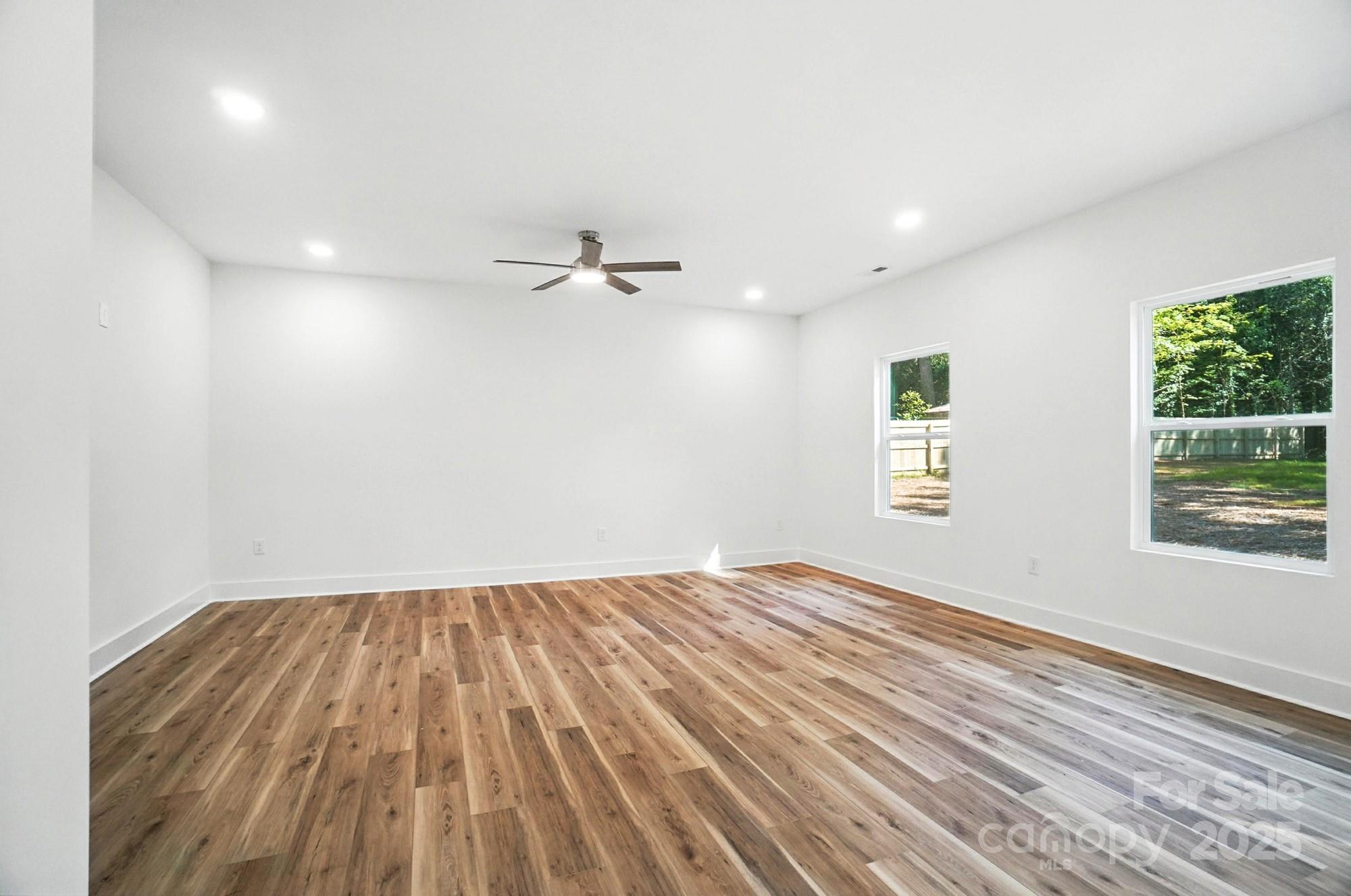 Vacant room with wood floors, a ceiling fan, and two windows overlooking trees.