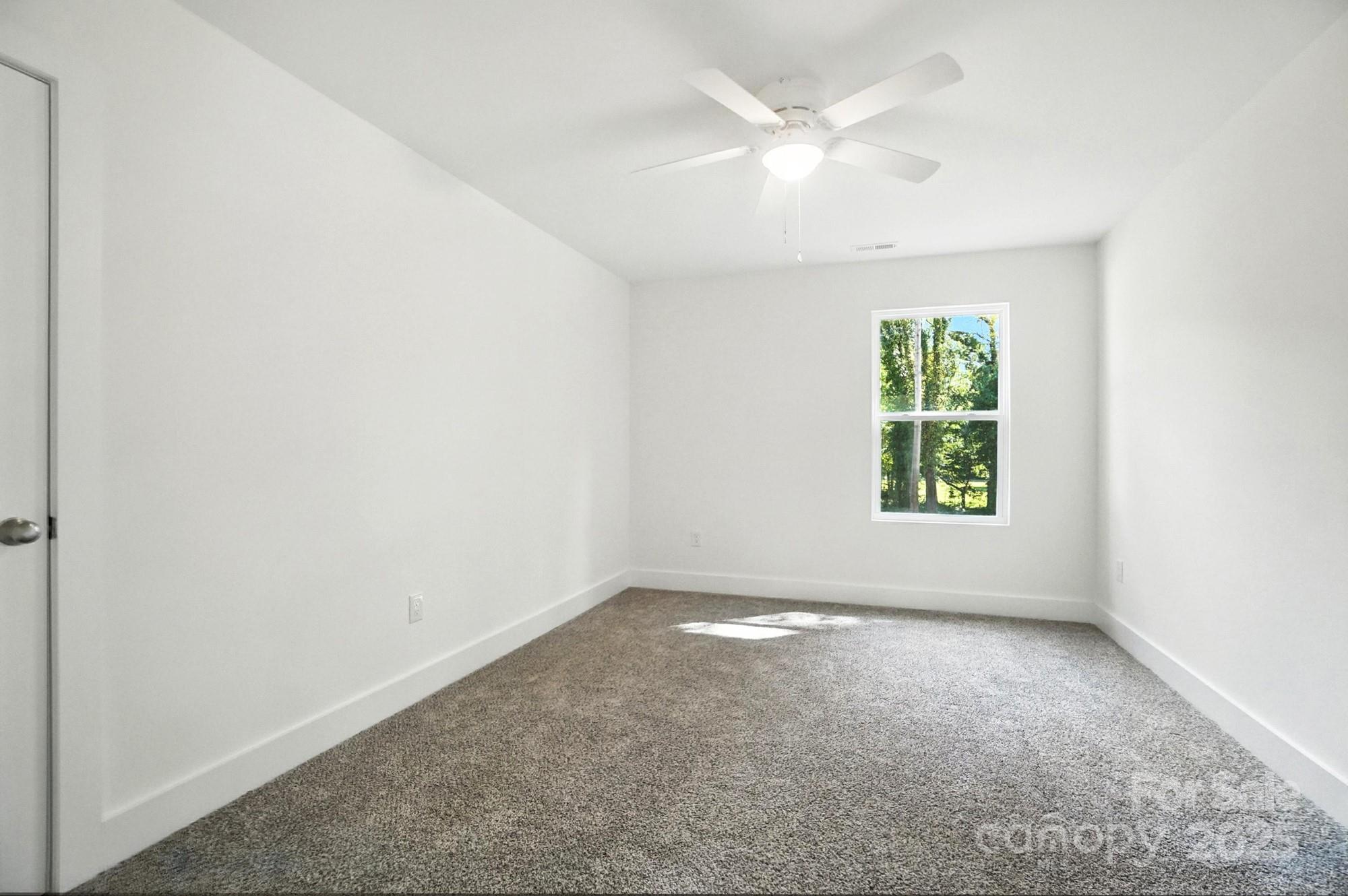 Empty bedroom with gray carpet, ceiling fan, and a window overlooking trees.