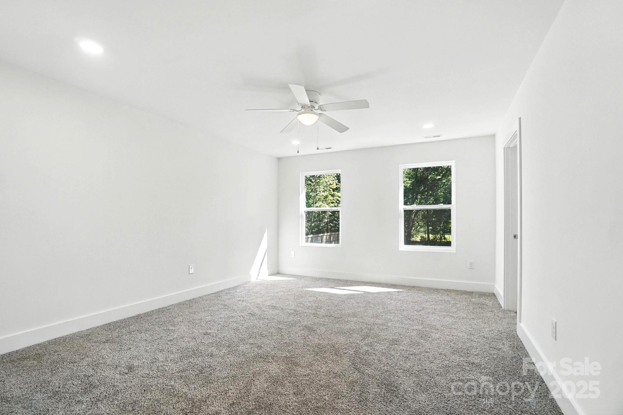 Vacant carpeted bedroom with a ceiling fan and two windows overlooking trees.