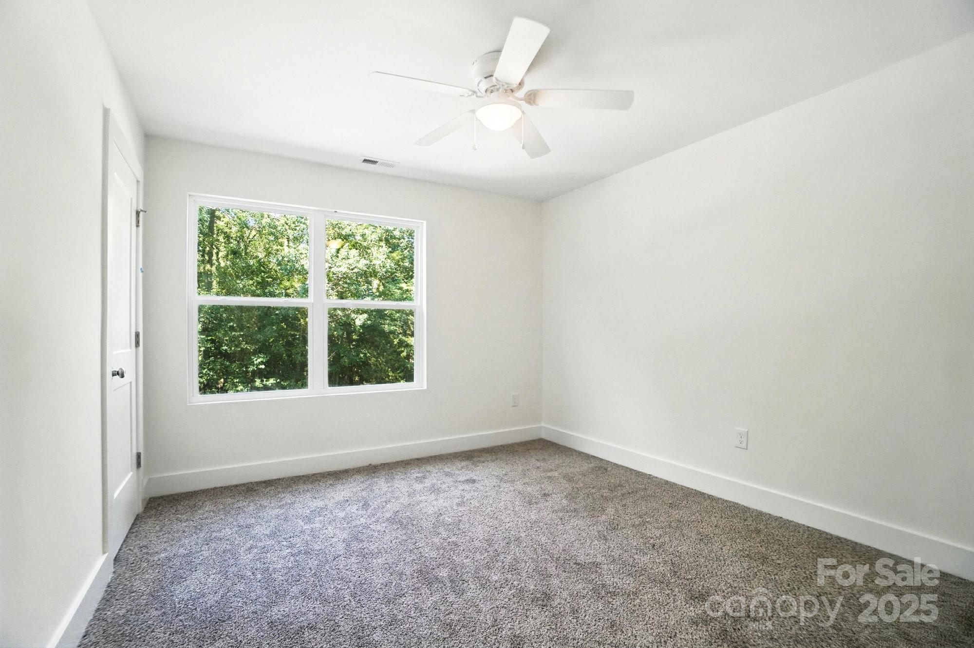 Empty bedroom with gray carpet, ceiling fan, and a large window overlooking trees.