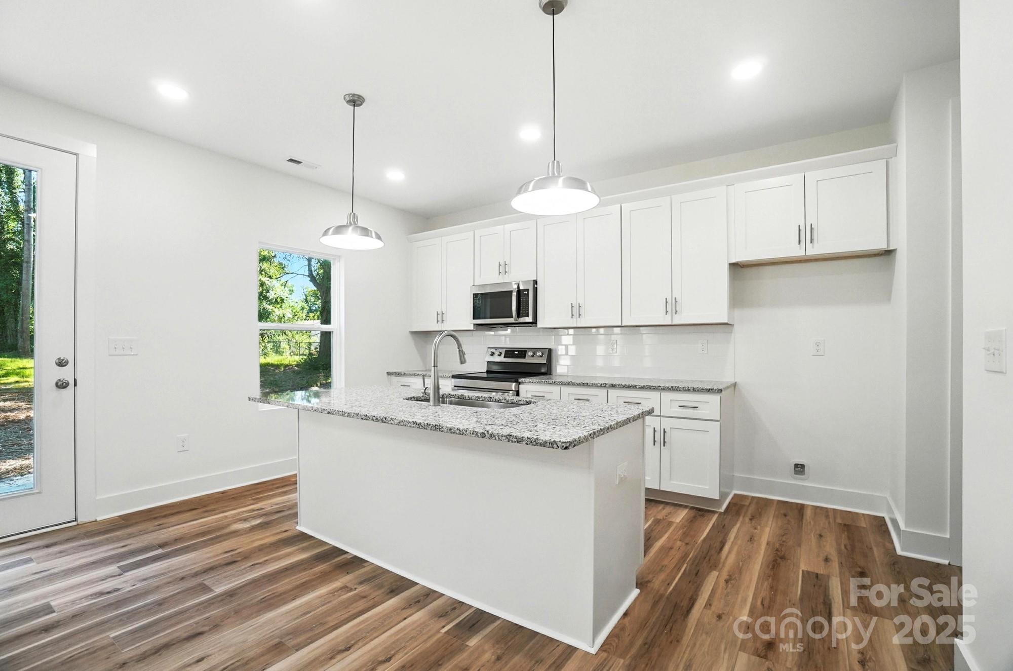Bright kitchen with white cabinets, stainless appliances, granite island, and wood-look floors.