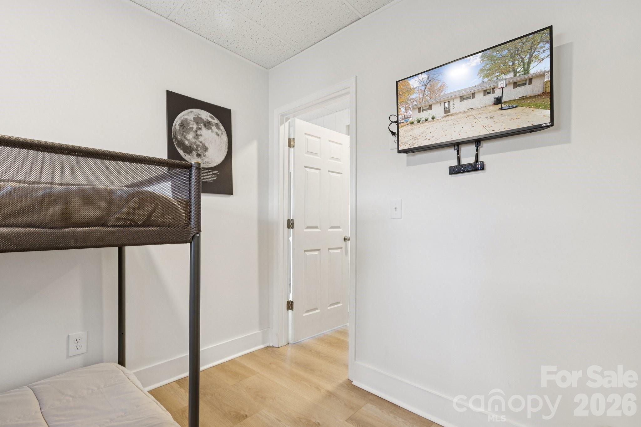 Bedroom with a black metal bunk bed, light wood flooring, and a wall-mounted TV showing the home’s exterior.