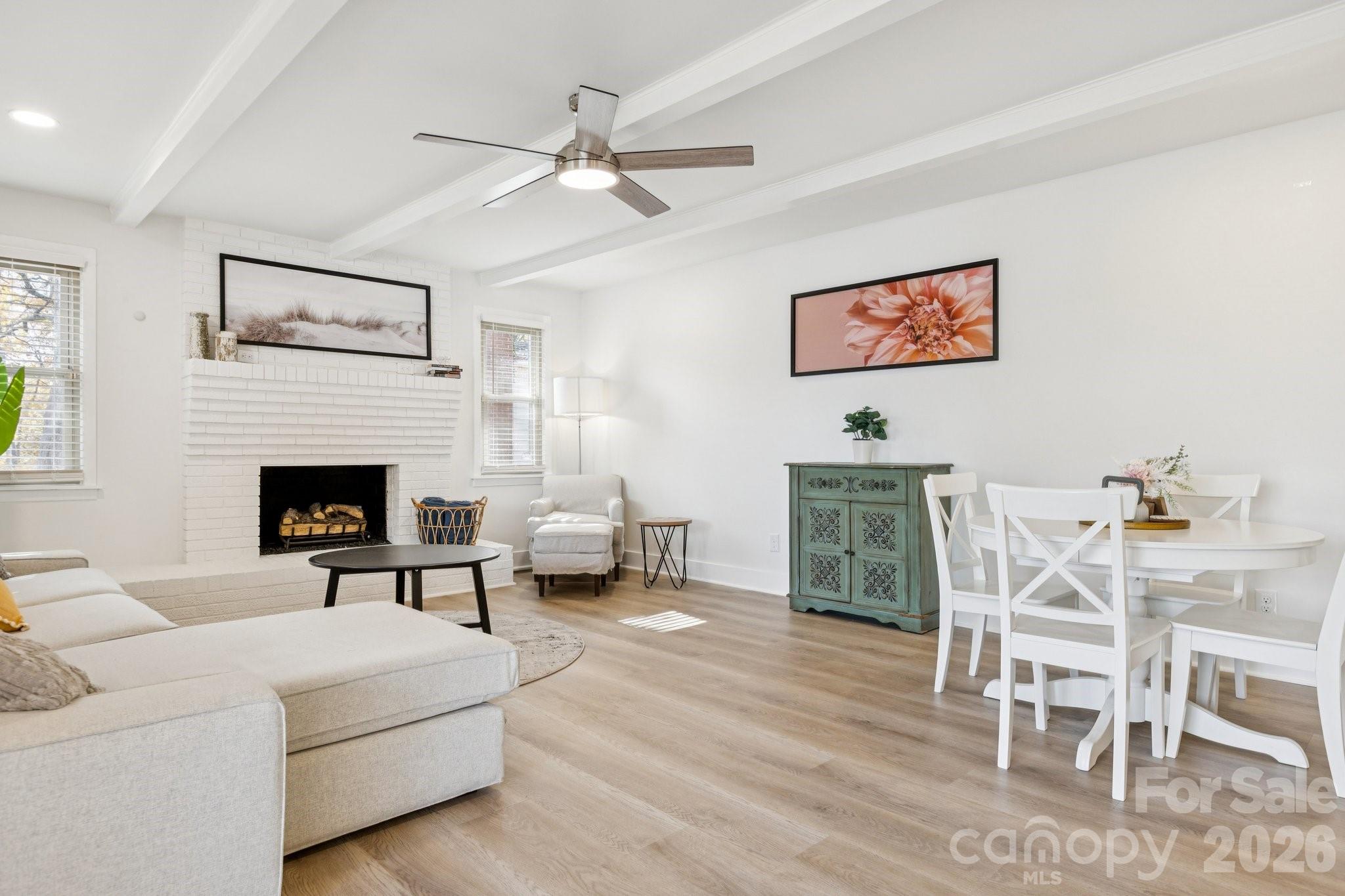 Living and dining room with white brick fireplace, exposed ceiling beams, and light wood floors.