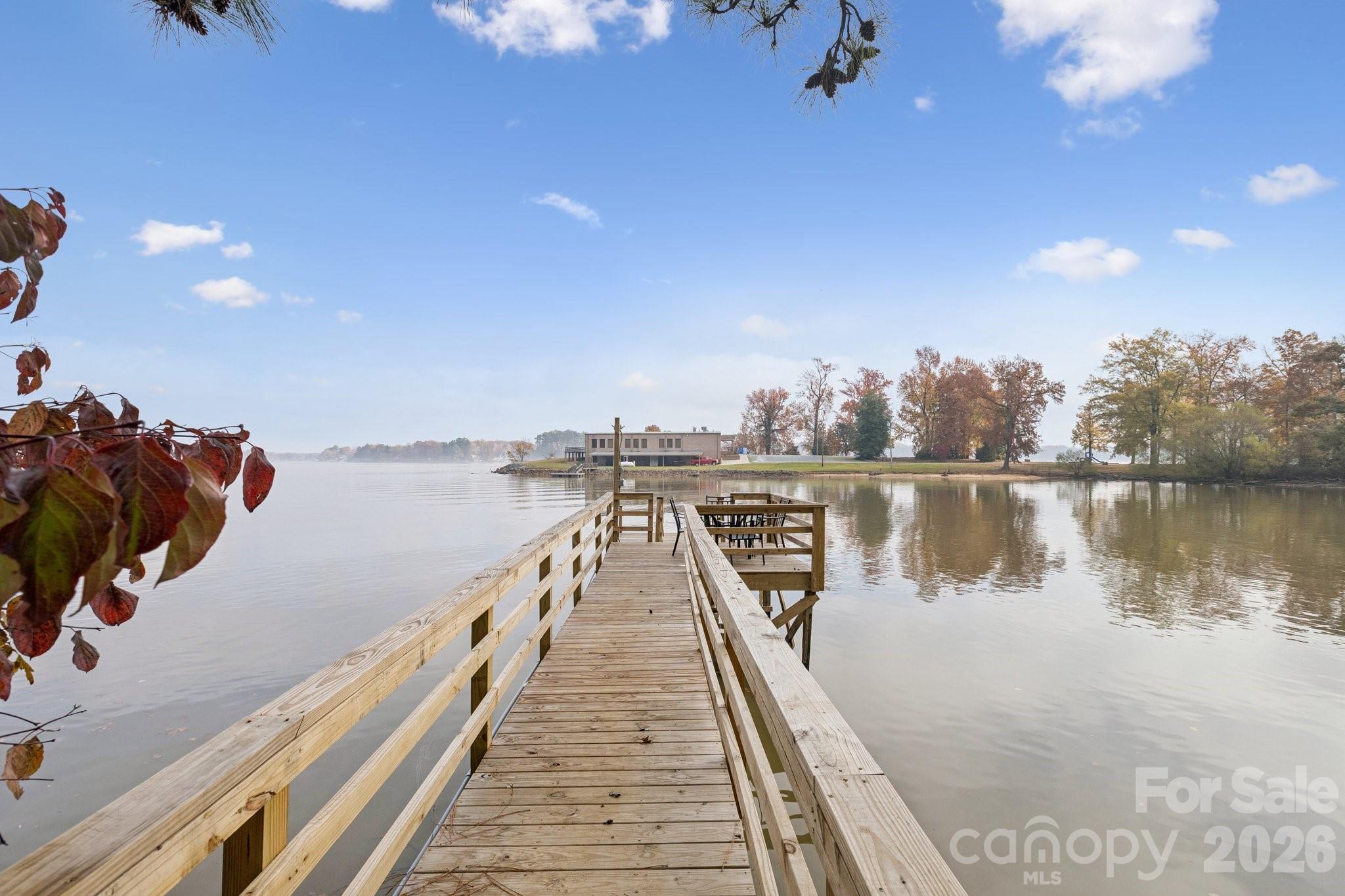 Wooden dock extending over calm water toward a distant building and autumn trees.