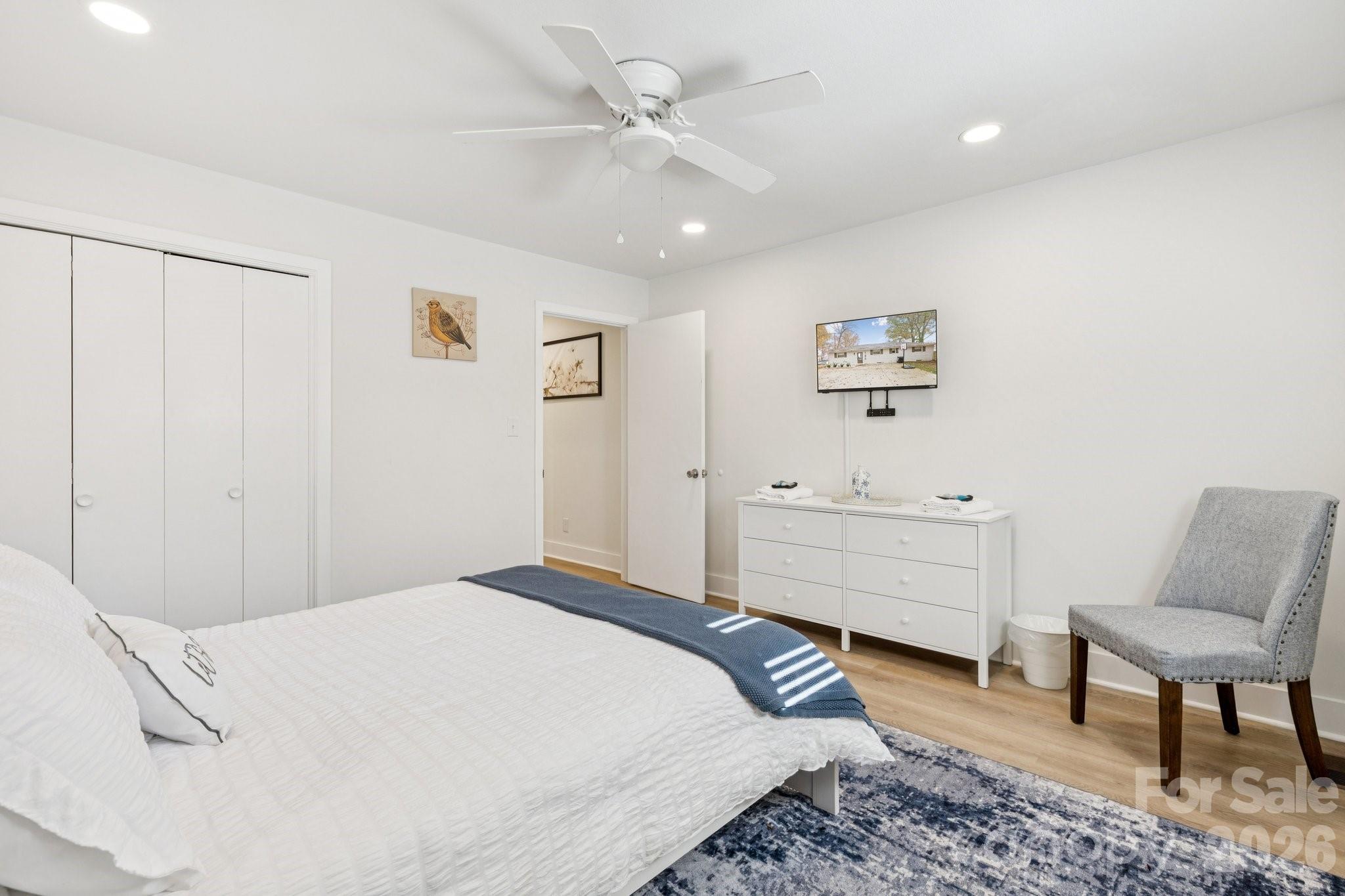 Staged bedroom with a white bed, blue throw, wall-mounted TV, and gray accent chair.