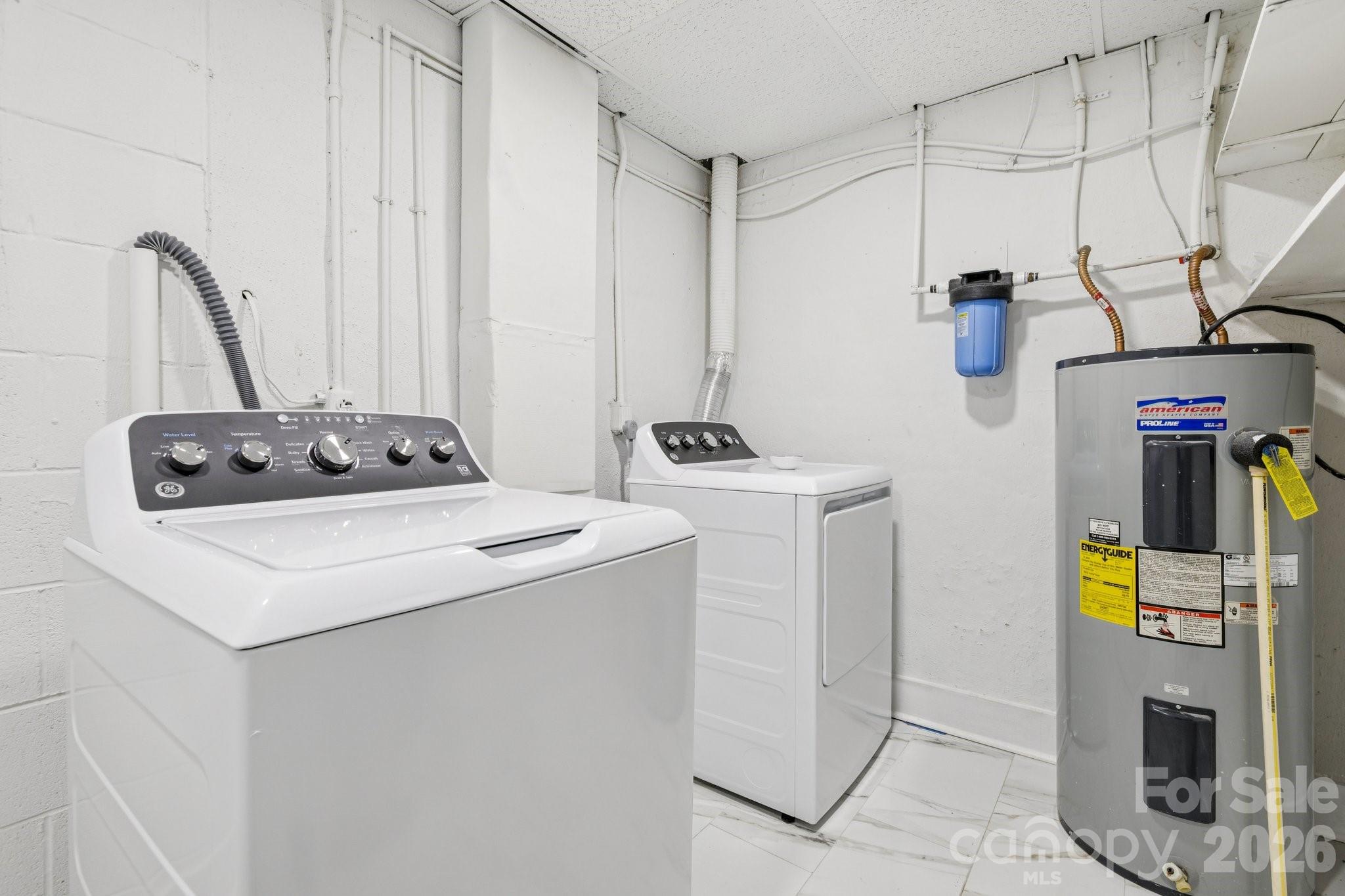 Laundry room with a top-loading washer, dryer, water heater, and exposed utility pipes.