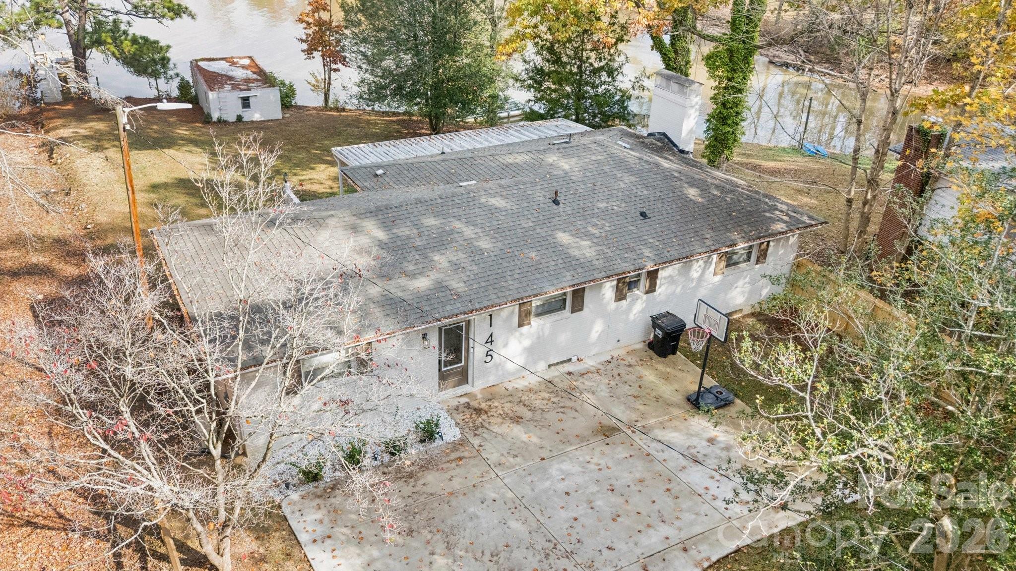 Aerial view of a single-story home beside the water, with a broad roof and basketball hoop by the driveway.