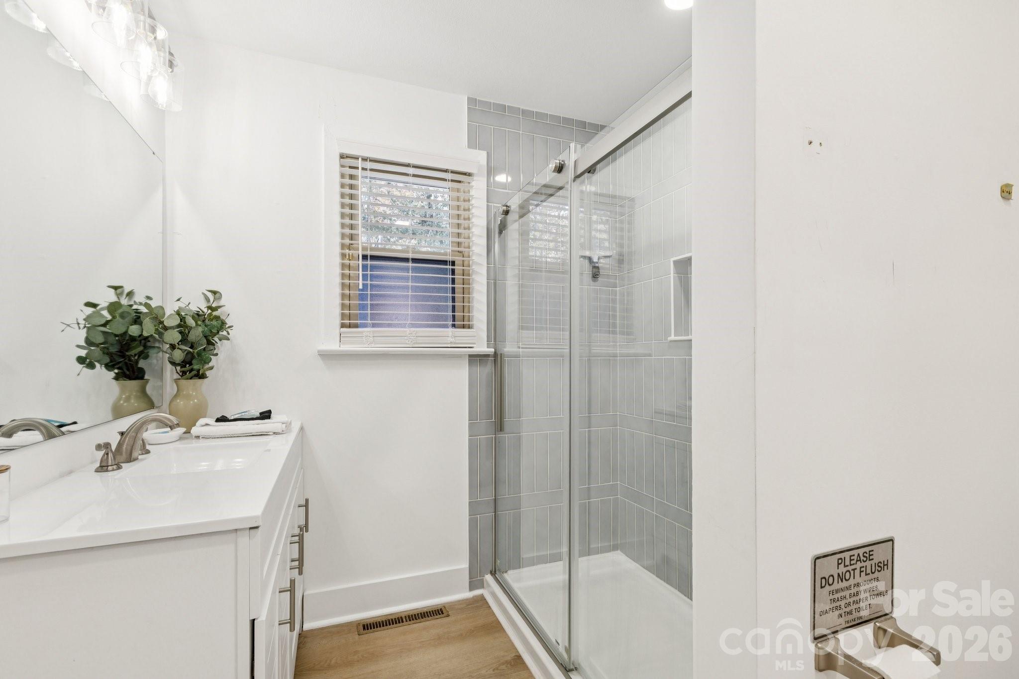 Bathroom with gray-tiled glass shower, white vanity, and a window with blinds.