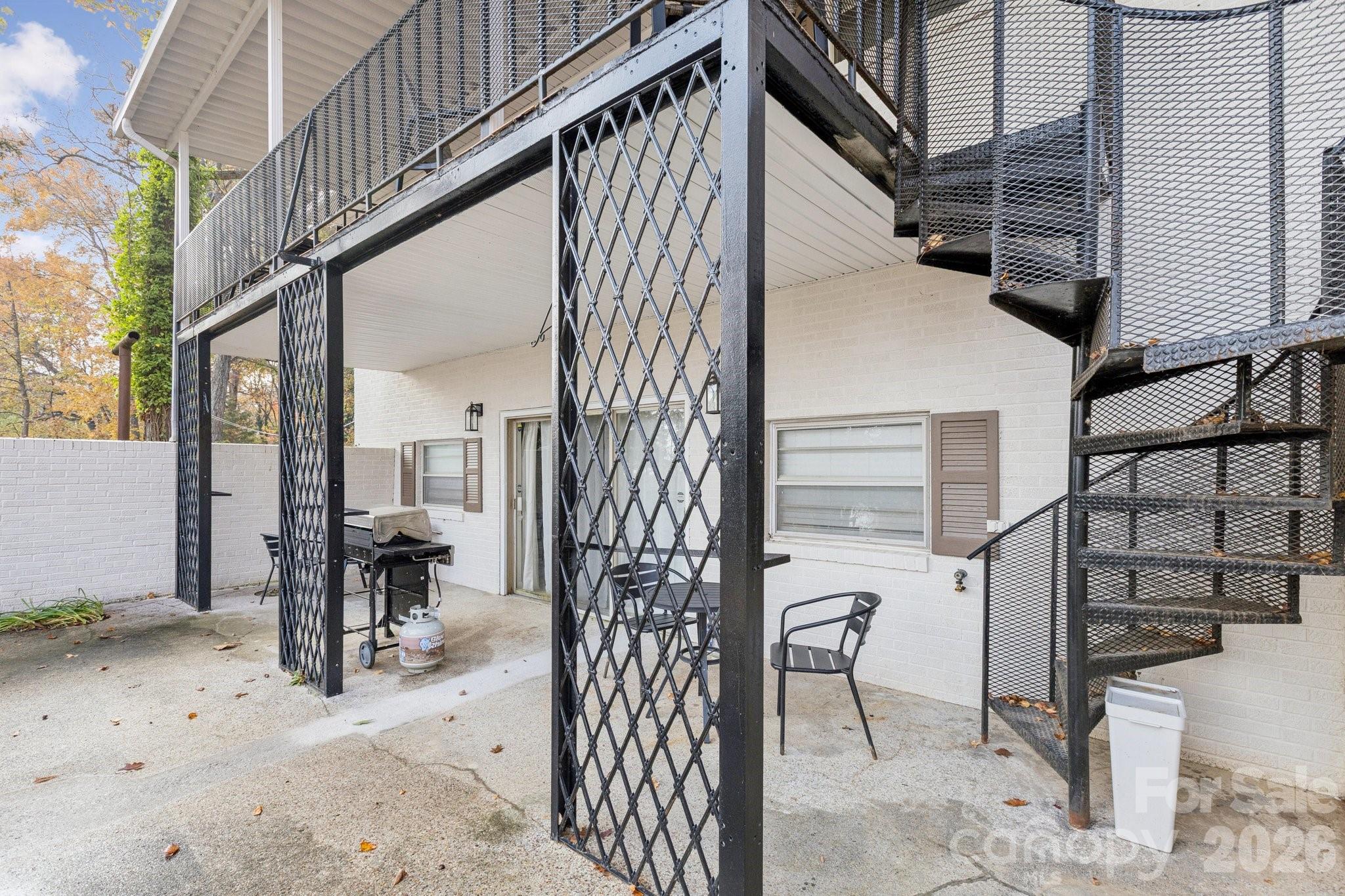 Patio beneath an elevated deck, with a black metal spiral staircase and grill beside a white brick home.