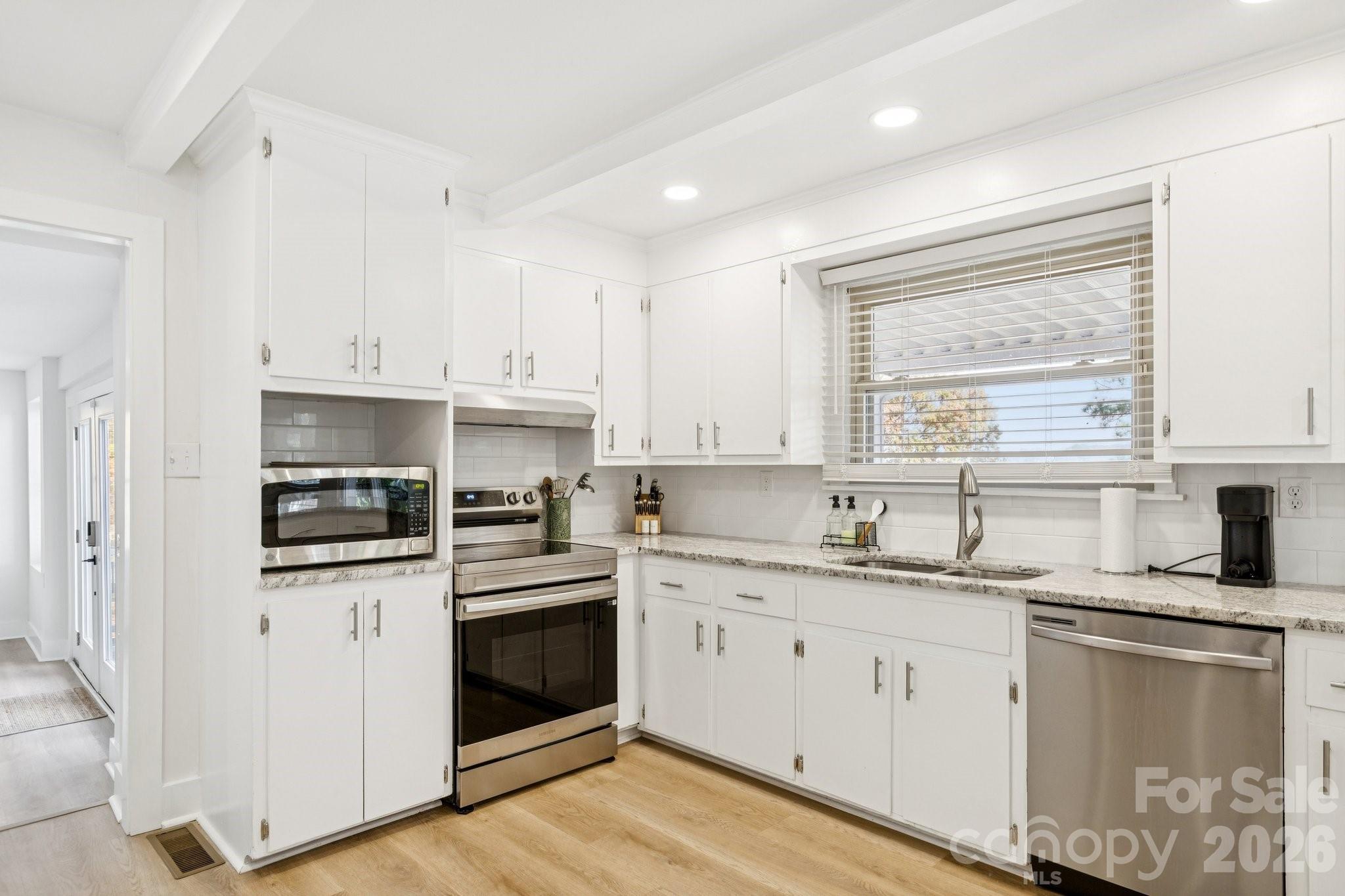 Bright white kitchen with stainless steel appliances, light wood floors, and a window above the sink.