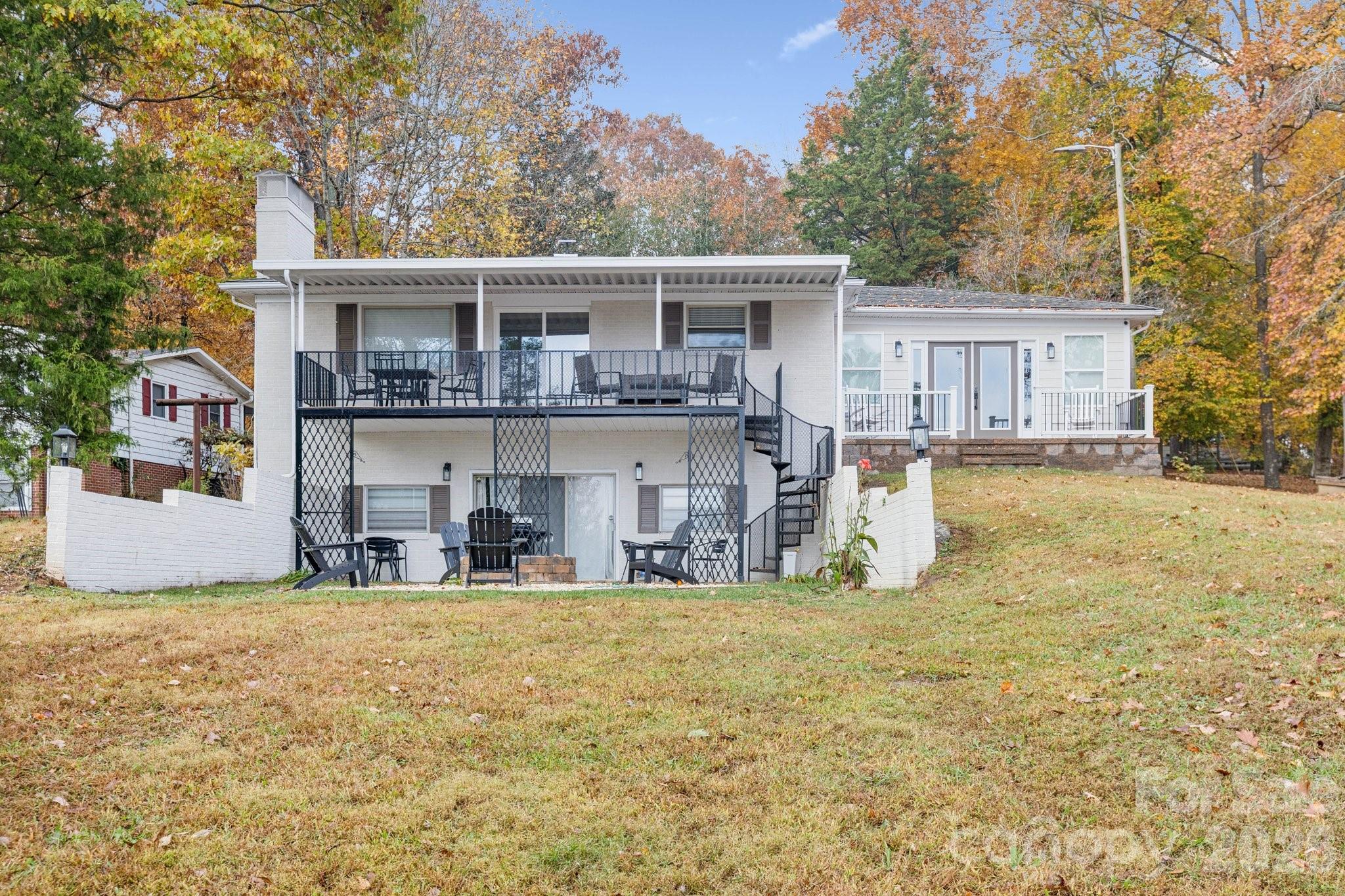 Rear of a white two-level house with covered deck, exterior stairs, and patio seating among autumn trees.