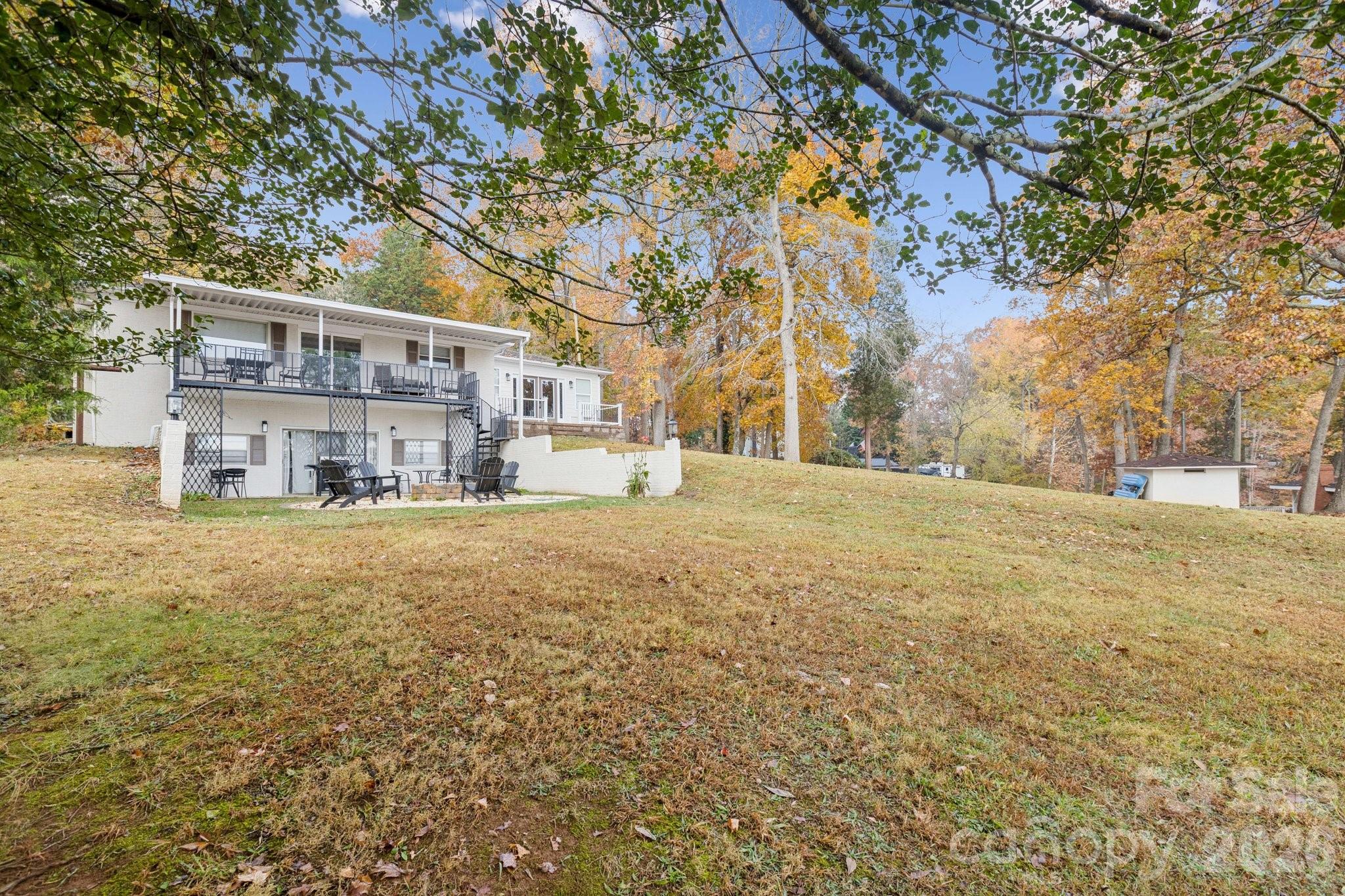 Rear of a white home with an elevated deck, patio seating, and a broad lawn beneath autumn trees.