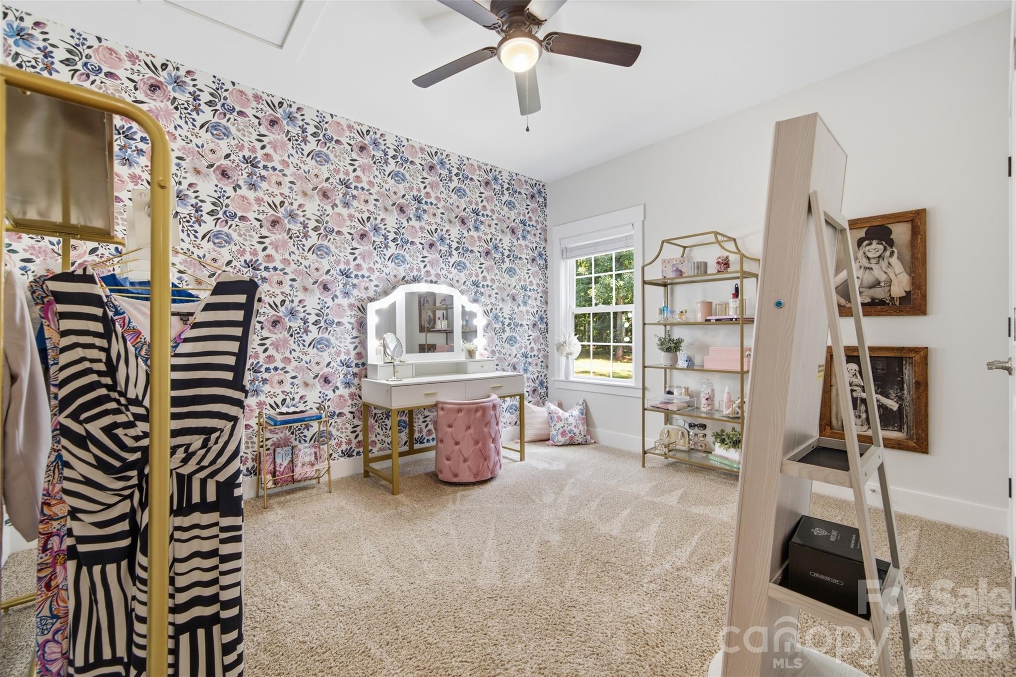 Dressing room with floral wallpaper, a white vanity, pink stool, clothing rack, and open shelves.