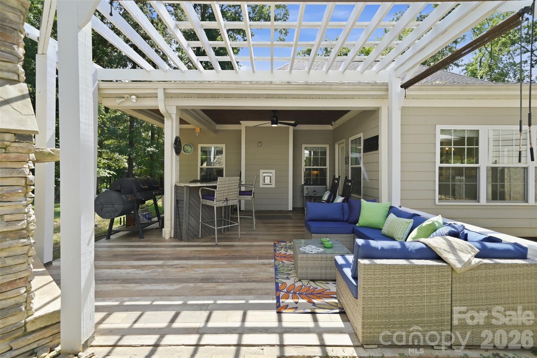 Covered patio with a white pergola, blue sectional seating, bar, and smoker grill.
