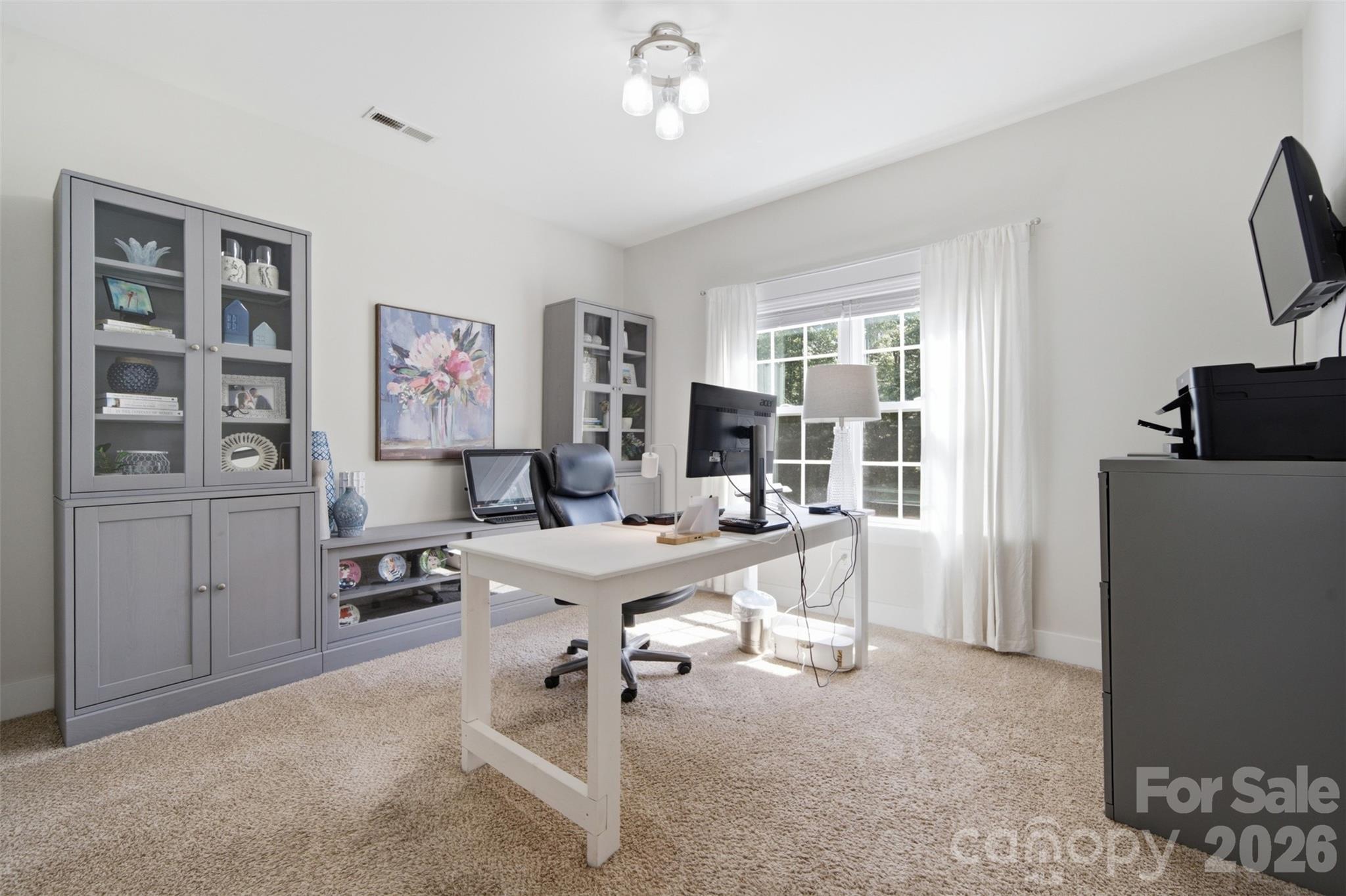 Furnished home office with a white desk, gray cabinets, and a sunlit window.