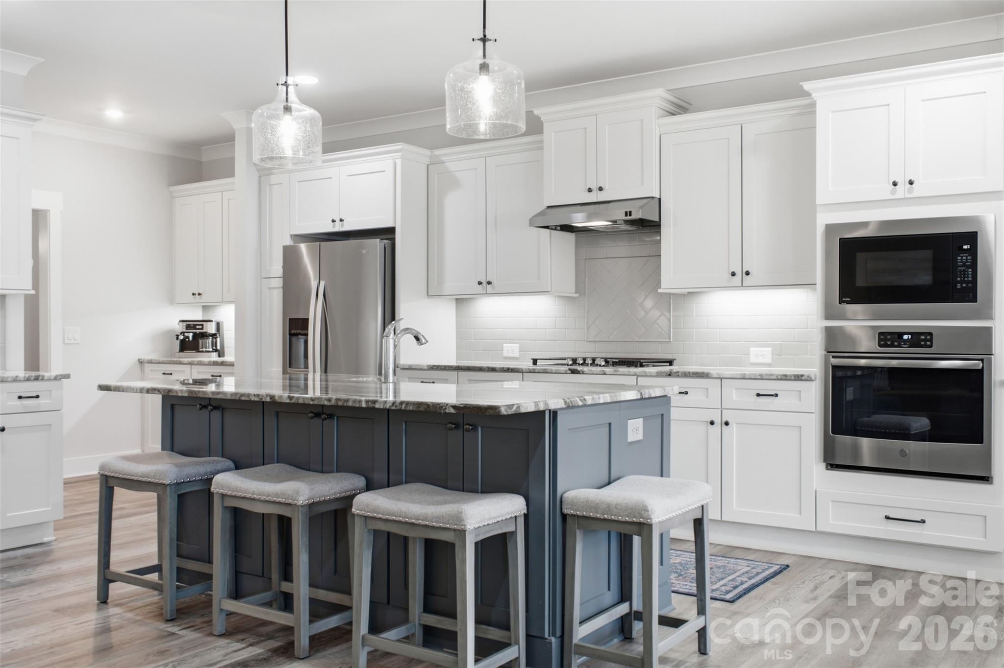 Kitchen with a dark blue-gray island, marble countertop, four stools, and white cabinets with stainless steel appliances.