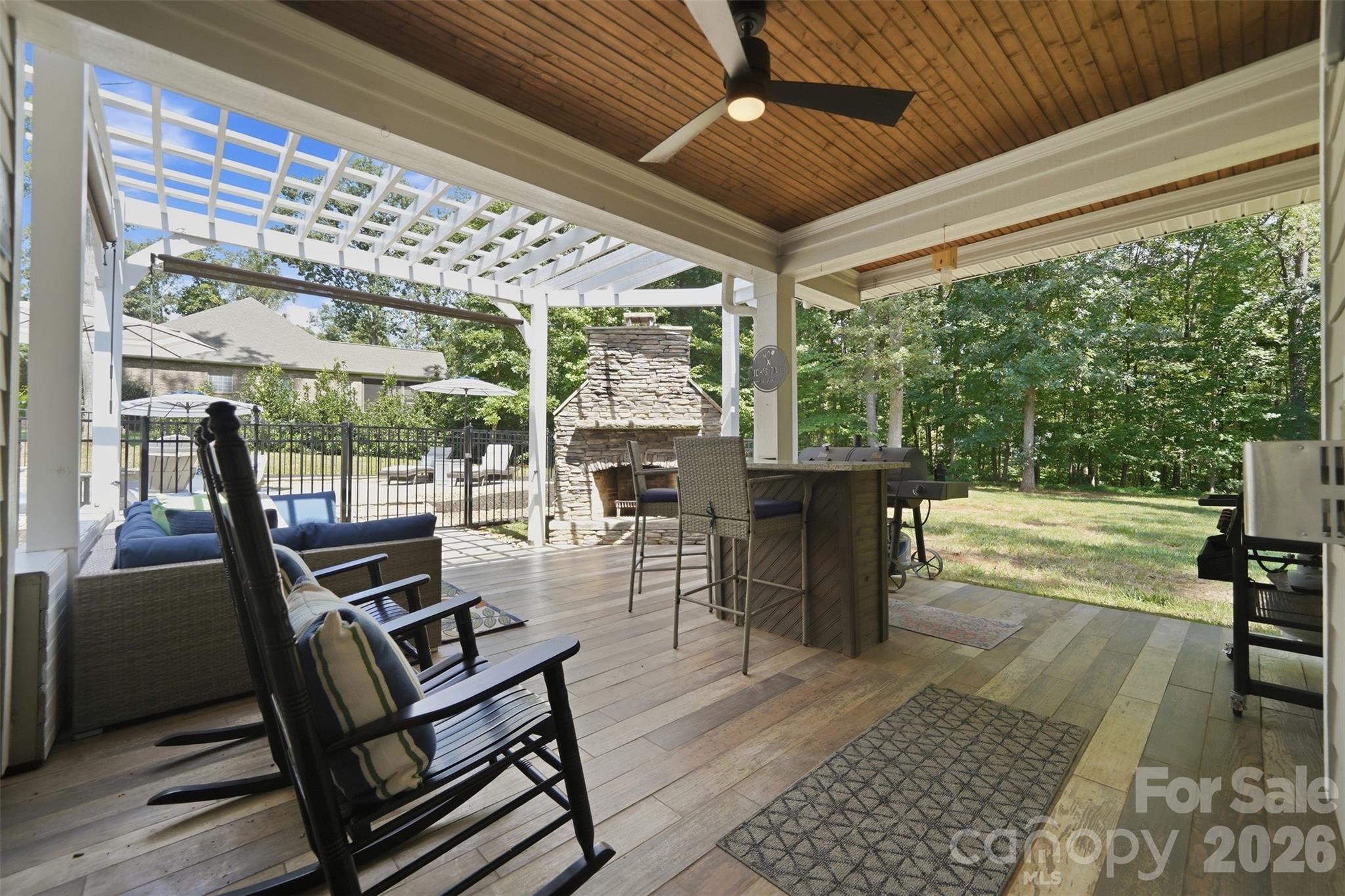 Covered patio with stone fireplace, bar seating, grill, and lounge chairs overlooking a wooded backyard.