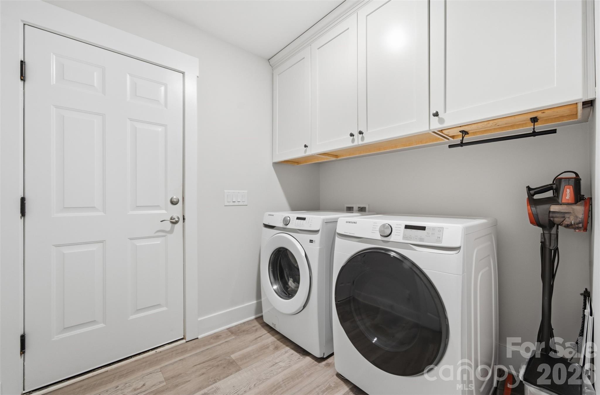 Laundry room with white front-loading washer and dryer beneath cabinets and a wood shelf.