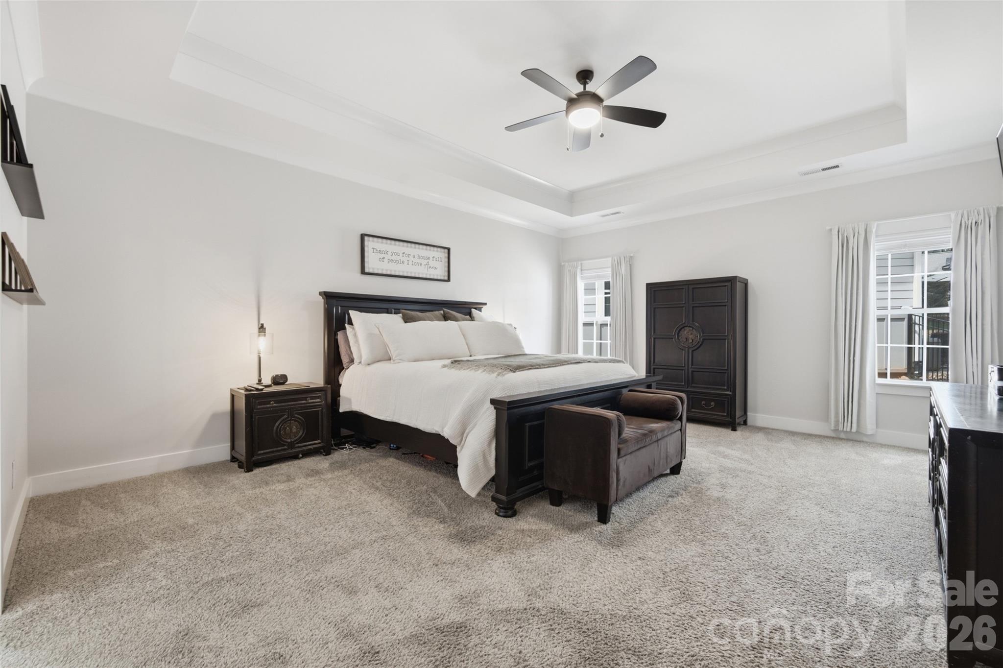 Furnished bedroom with a dark wood bed, white bedding, carpet, and a tray ceiling with ceiling fan.