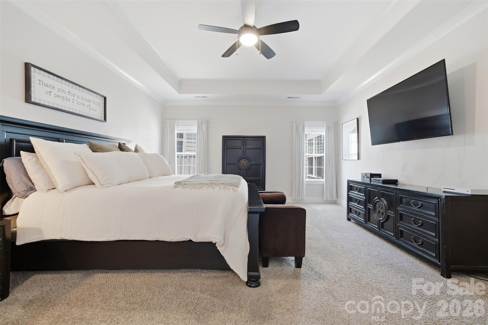 Furnished bedroom with a large white bed, tray ceiling, ceiling fan, and wall-mounted television.