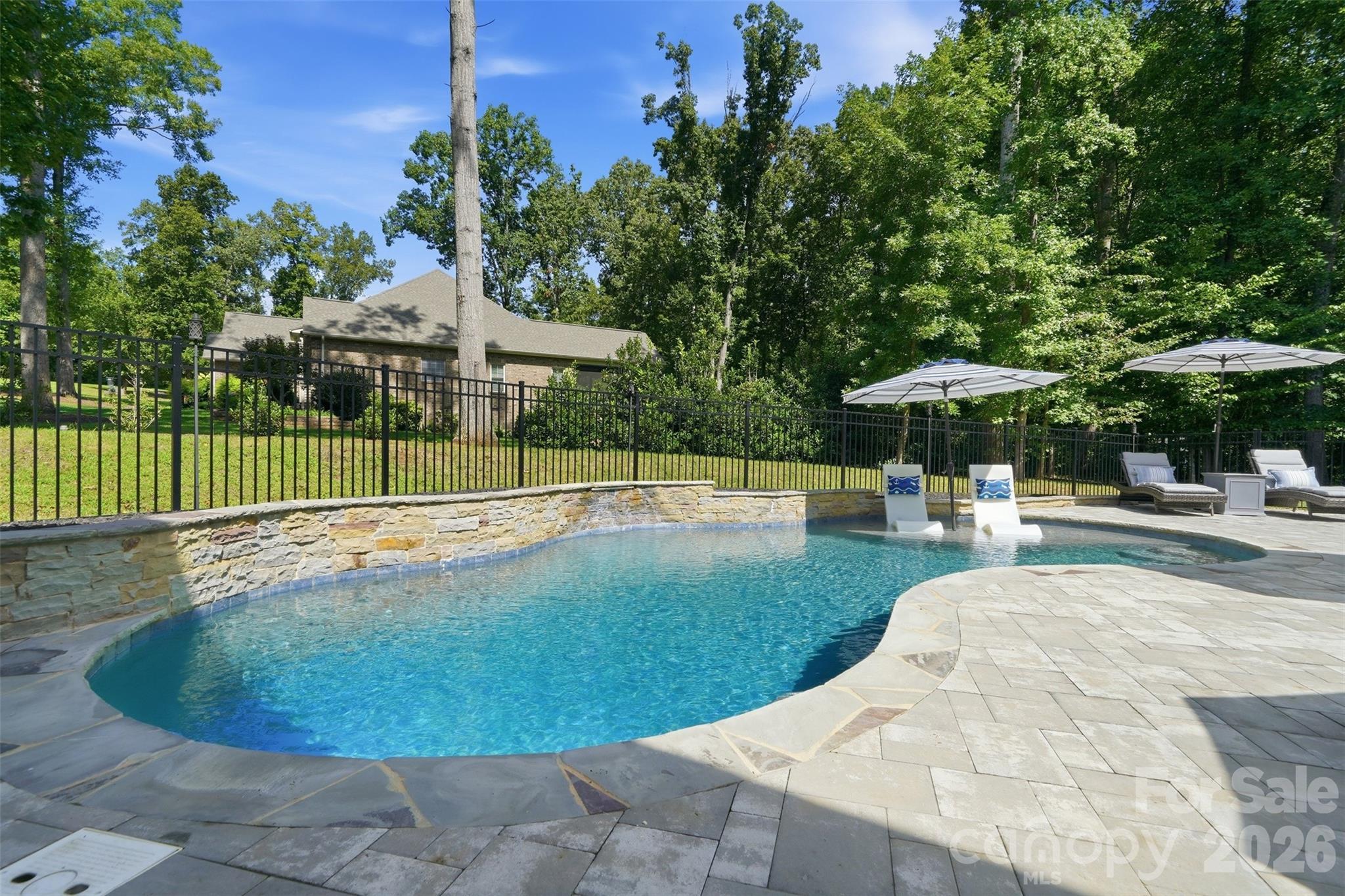 Curved backyard pool with in-water loungers, patio umbrellas, and a wooded setting.