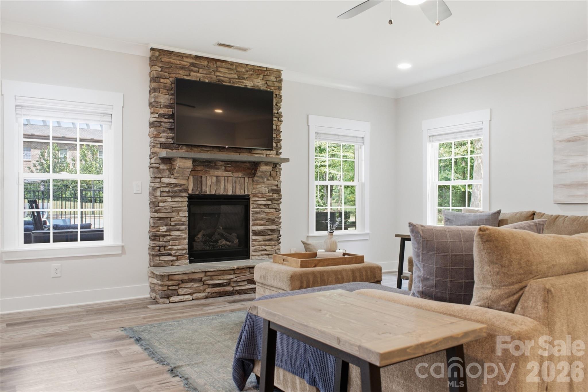 Staged living room with a stone fireplace, mounted television, and large windows.