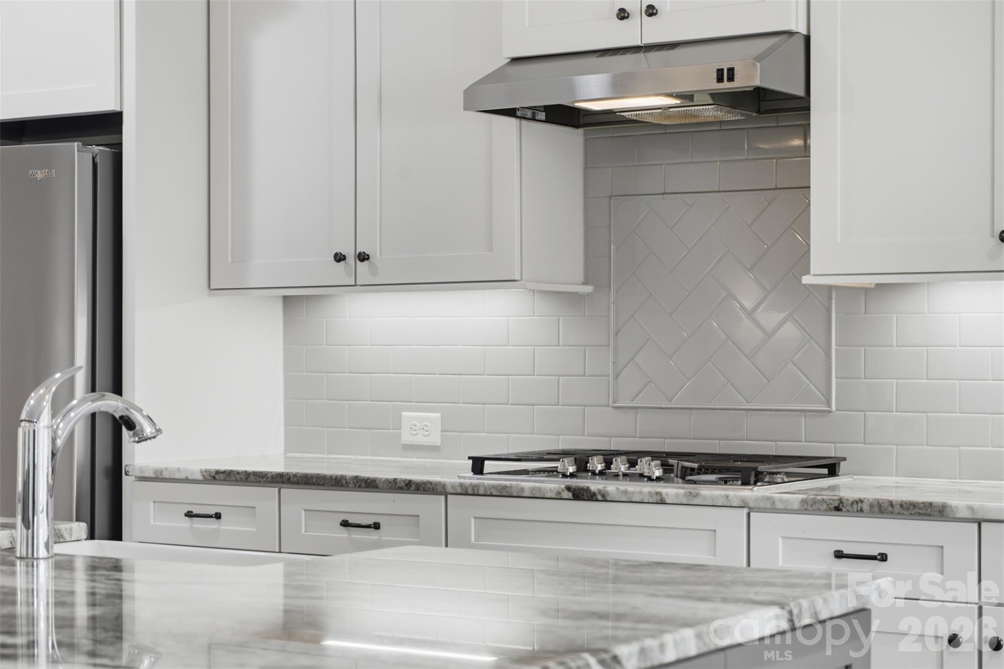 White kitchen with herringbone tile accent, gas cooktop, stainless range hood, and marble-look countertops.