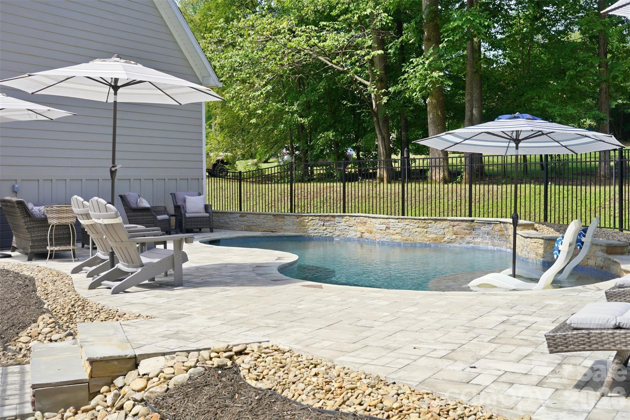 Backyard pool with a stone-paver patio, shaded lounge chairs, and a black metal fence.