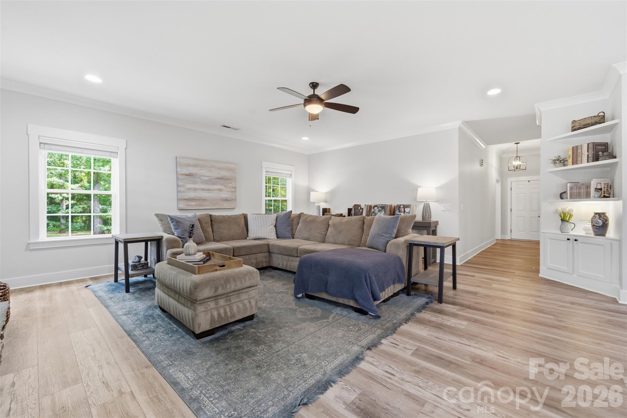 Staged living room with a sectional sofa, blue-gray rug, built-in shelves, and windows facing trees.