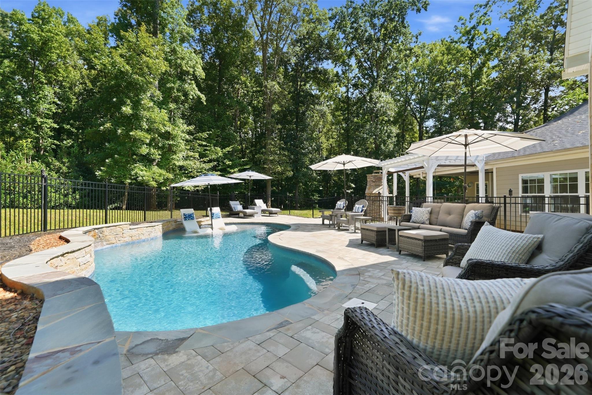 Curved backyard pool with in-pool loungers, patio seating, and a wooded backdrop.