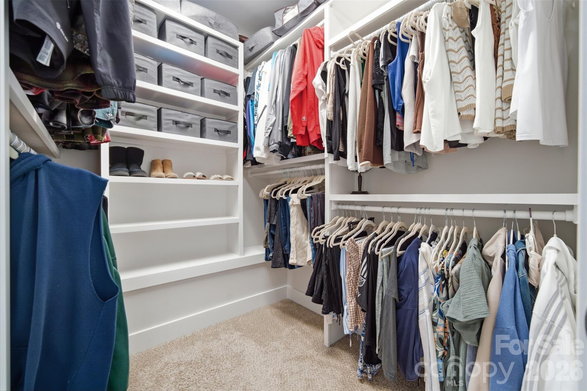 Walk-in closet with white built-in shelves, storage bins, shoe storage, and clothes hanging on two walls.