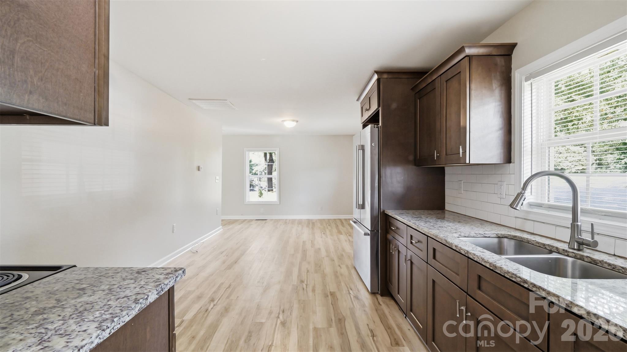 Kitchen with dark wood cabinets, speckled countertops, and a window above the sink, opening to an unfurnished room.