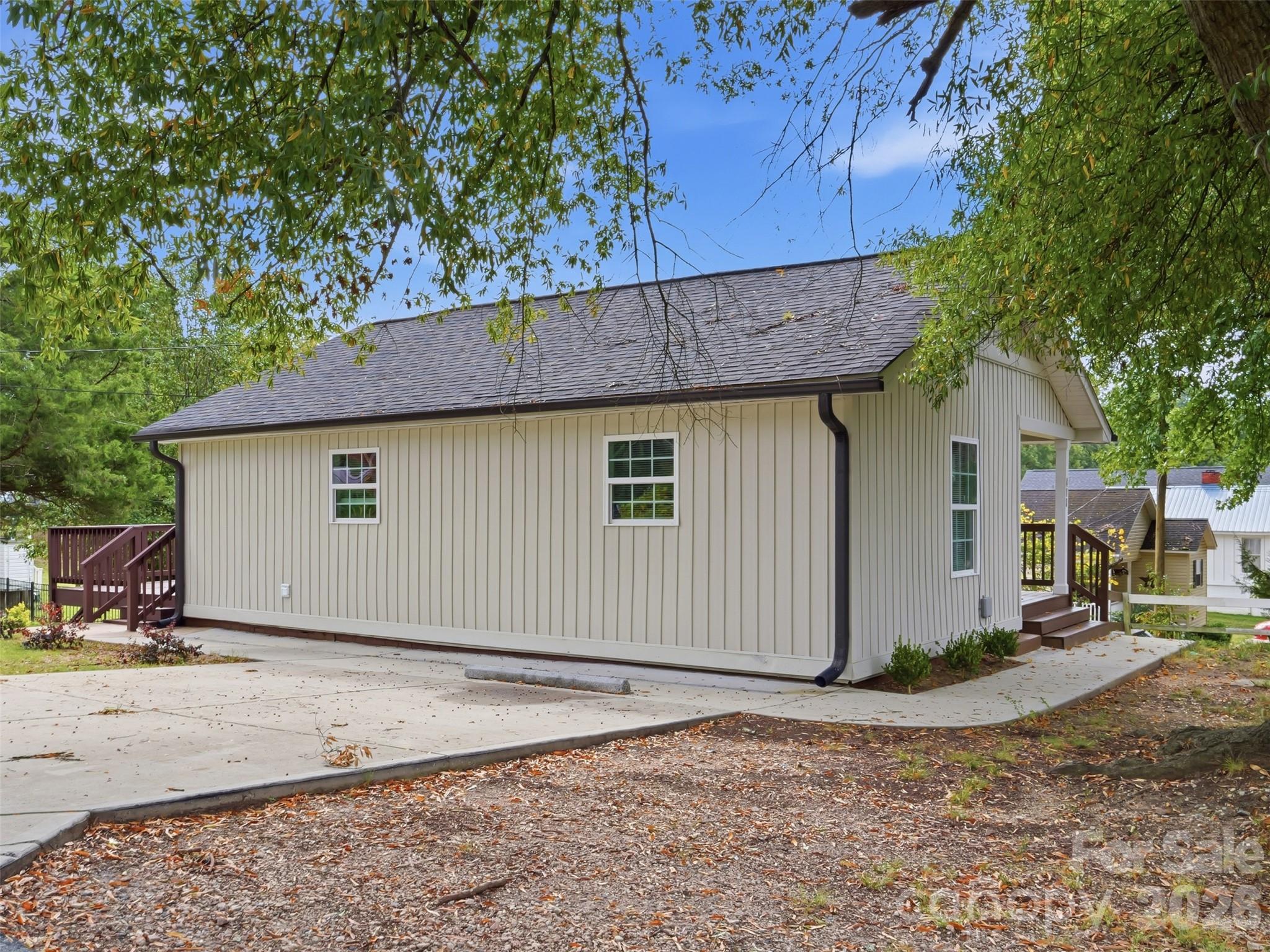 Light-sided house with a dark shingle roof, wooden deck stairs, and a broad concrete driveway.