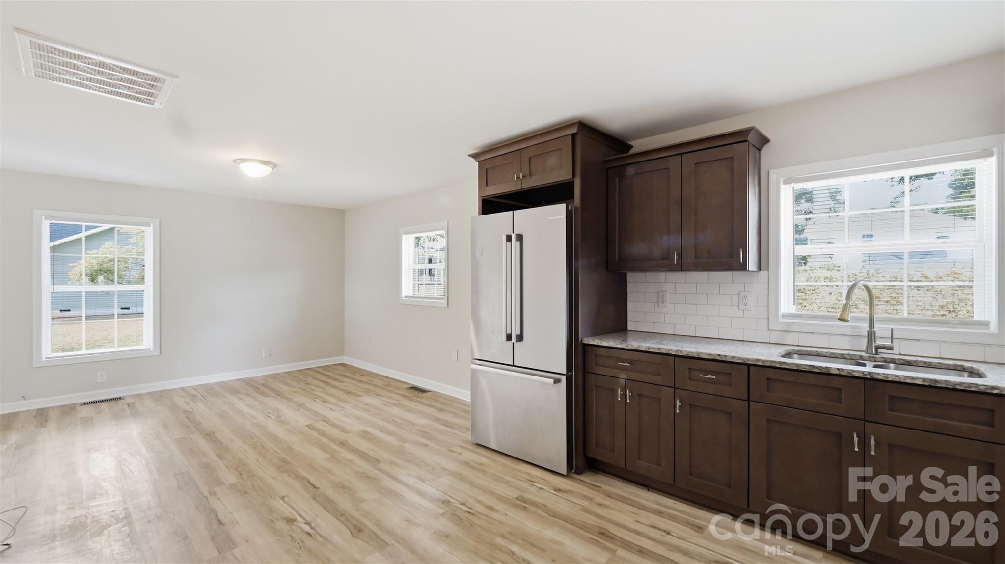 Vacant kitchen with dark cabinets, stainless steel refrigerator, subway tile backsplash, and light wood-look flooring.