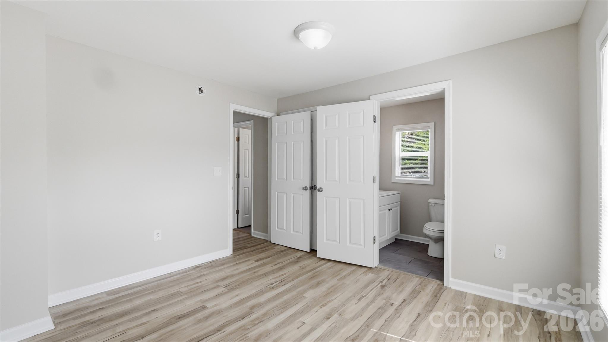Empty bedroom with wood-look flooring and open doors to a bathroom and hallway.