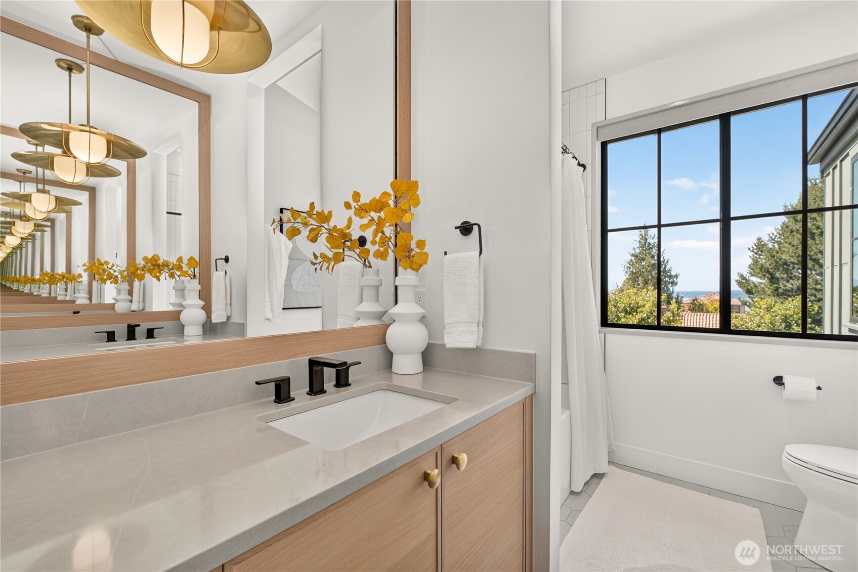 Bathroom with a light wood vanity, oversized mirrors, and a black-framed window overlooking water.