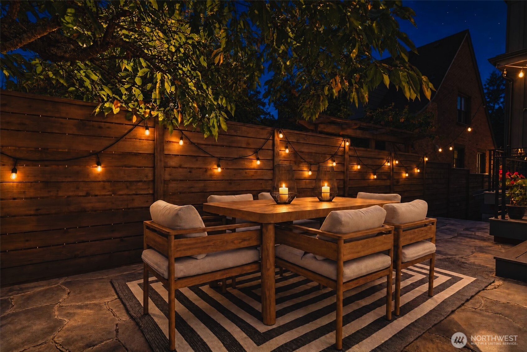 Outdoor dining patio with a wooden table and cushioned chairs beneath string lights along a privacy fence.