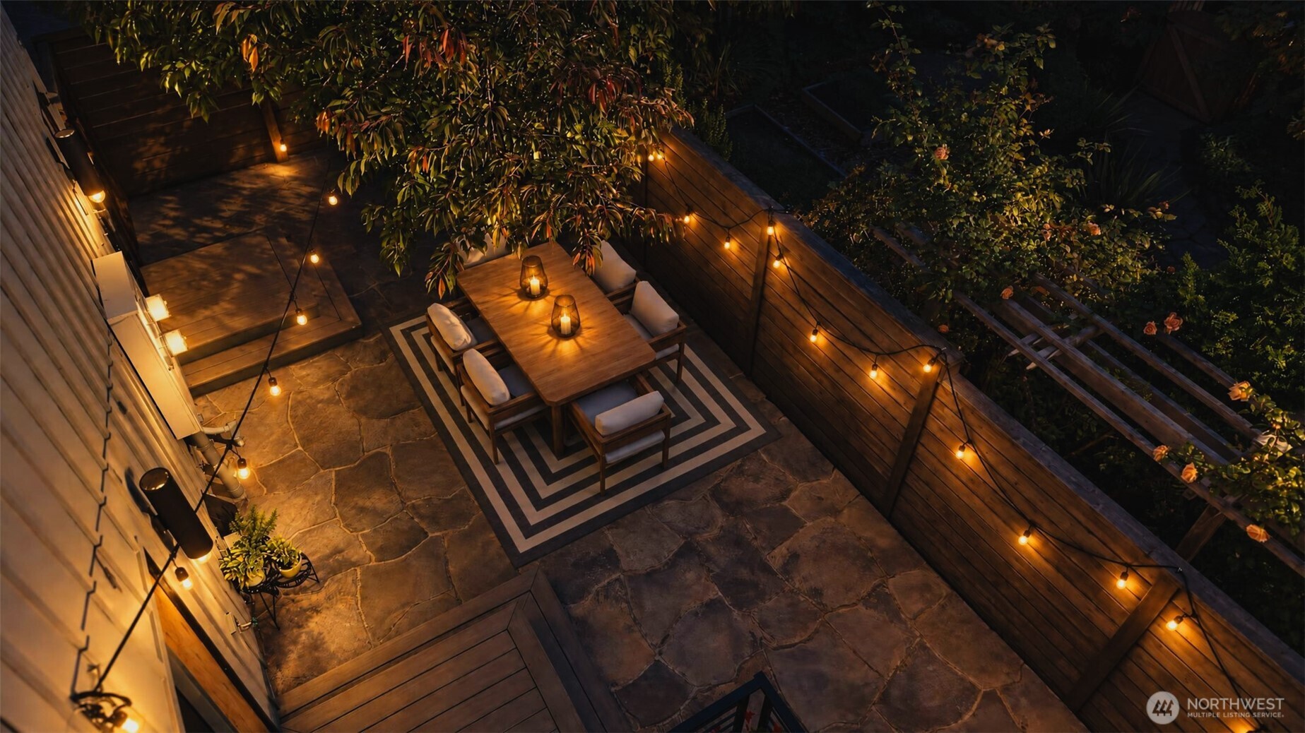 Stone patio dining area with cushioned chairs, glowing string lights, and a lush garden at night.