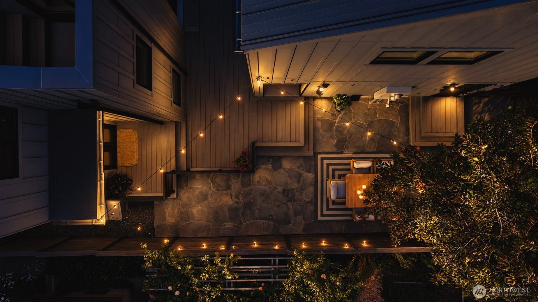 Overhead view of a stone patio with string lights, outdoor dining furniture, and surrounding landscaping.