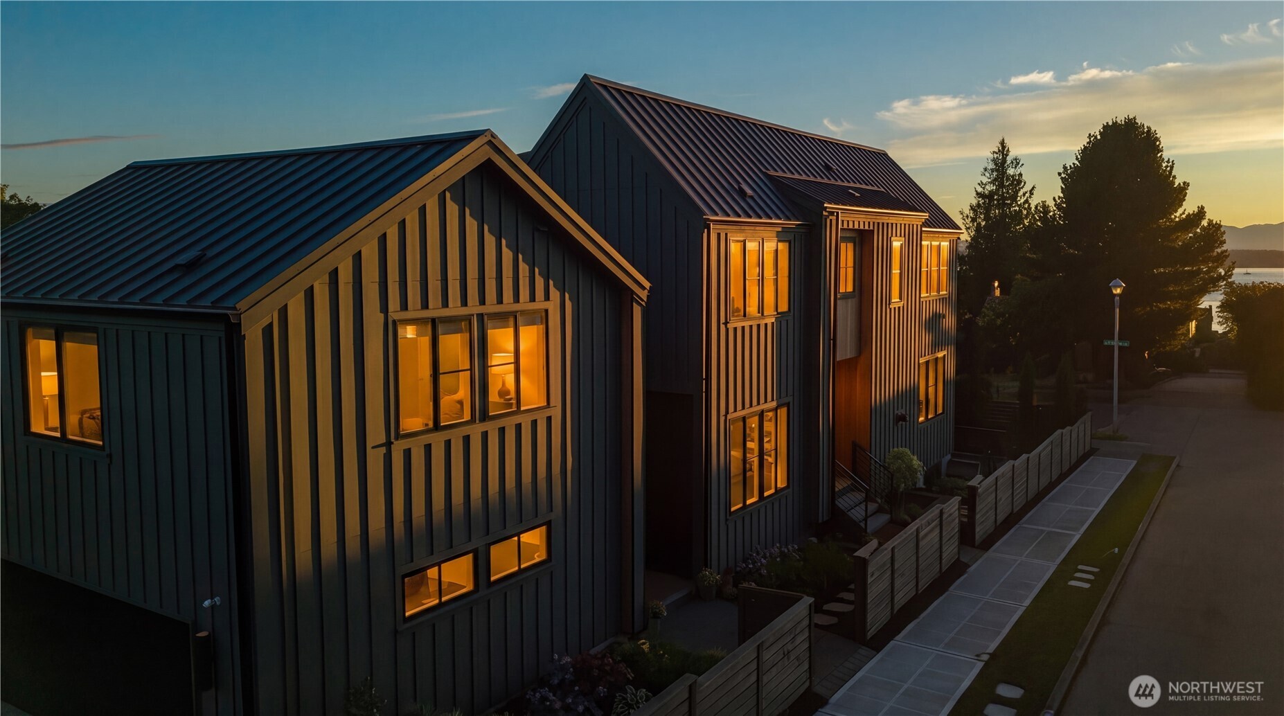Two modern dark-sided homes with metal roofs and glowing windows at dusk, with water and hills in the distance.