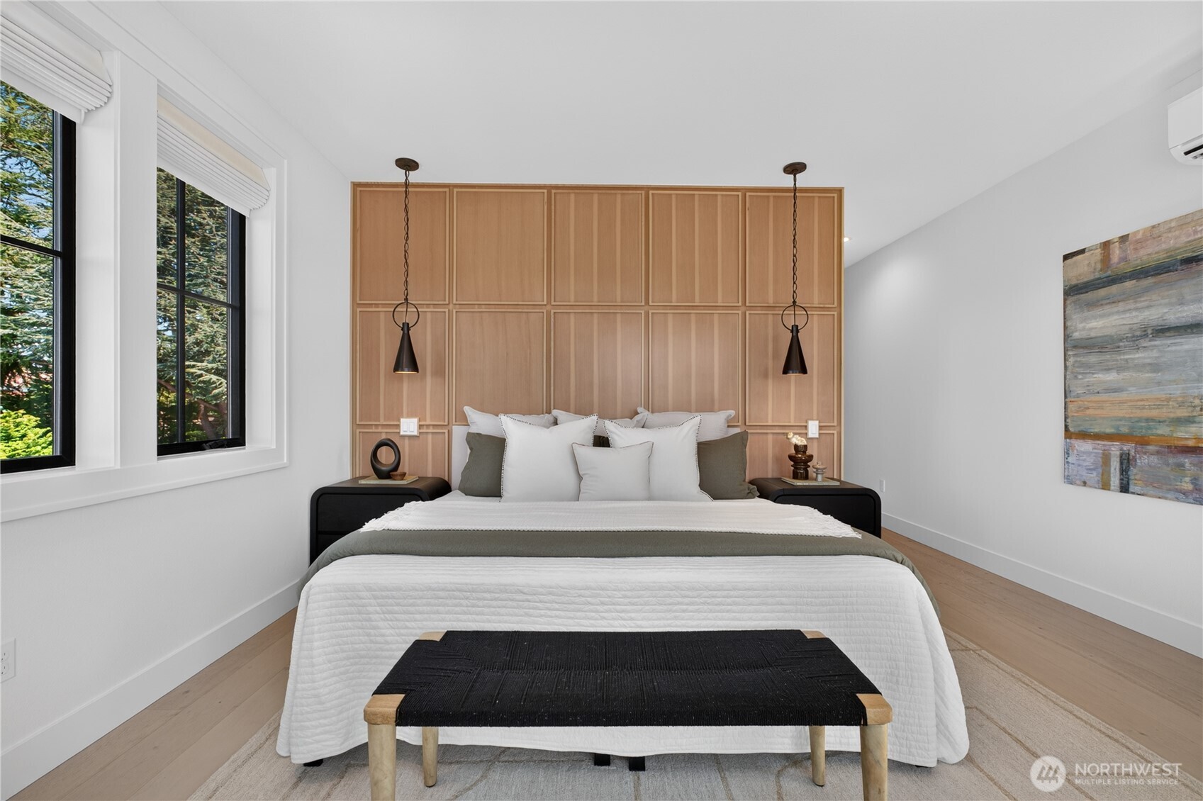 Staged bedroom with a paneled wood headboard, pendant lights, and large windows overlooking trees.