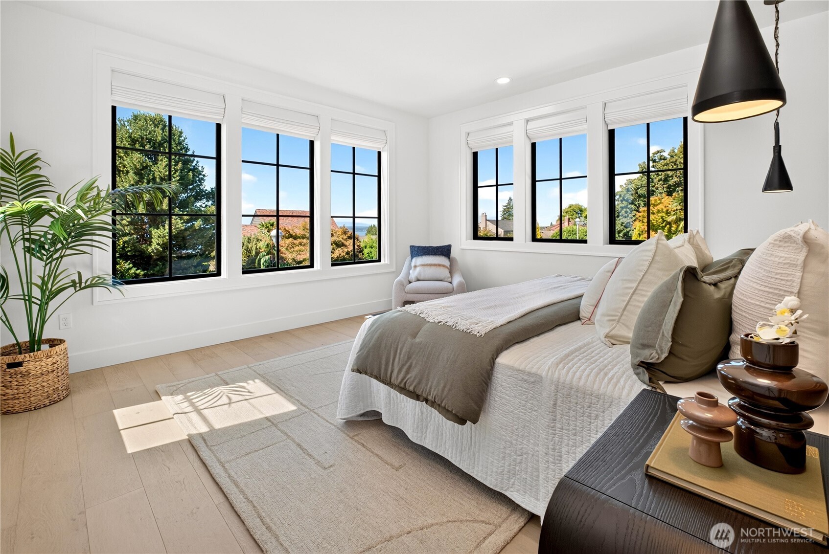 Bright staged bedroom with a large bed and tall windows overlooking leafy trees and rooftops.