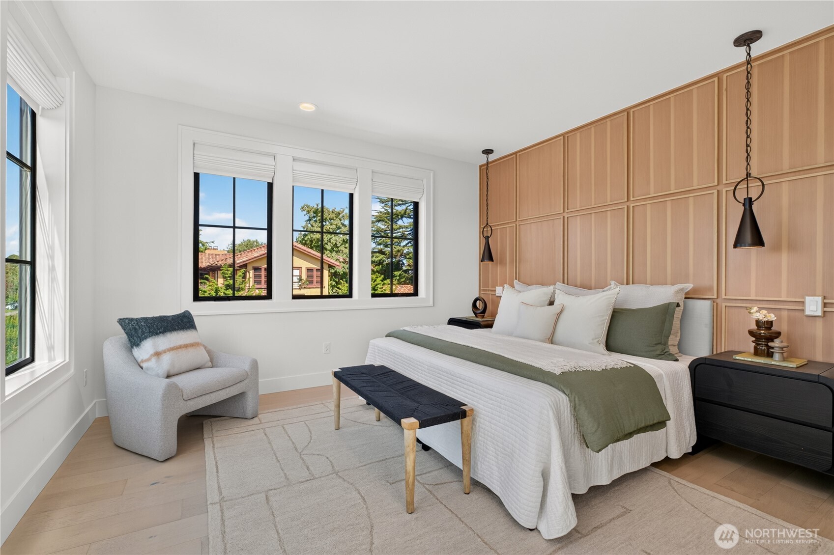 Staged bedroom with a wood-paneled accent wall, white bed, and large daylight-filled windows.