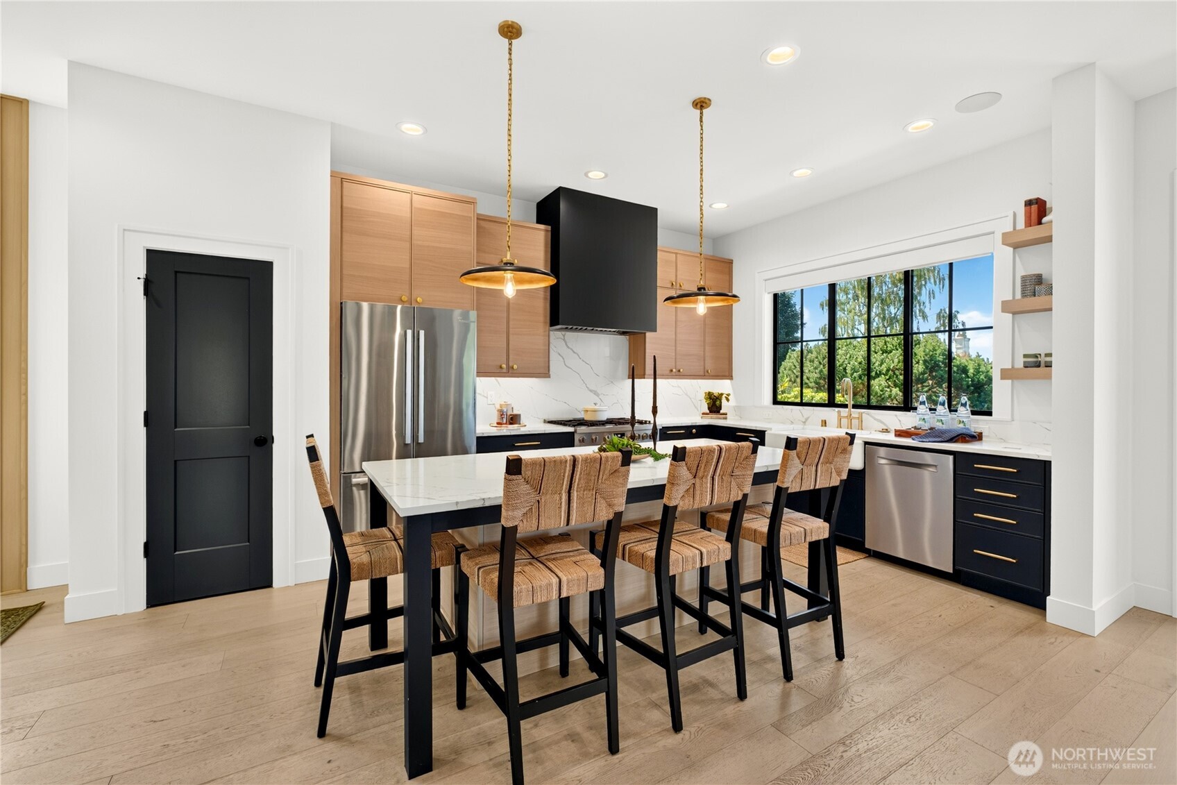 Kitchen with a large island, woven bar stools, light wood cabinets, and black-framed windows.