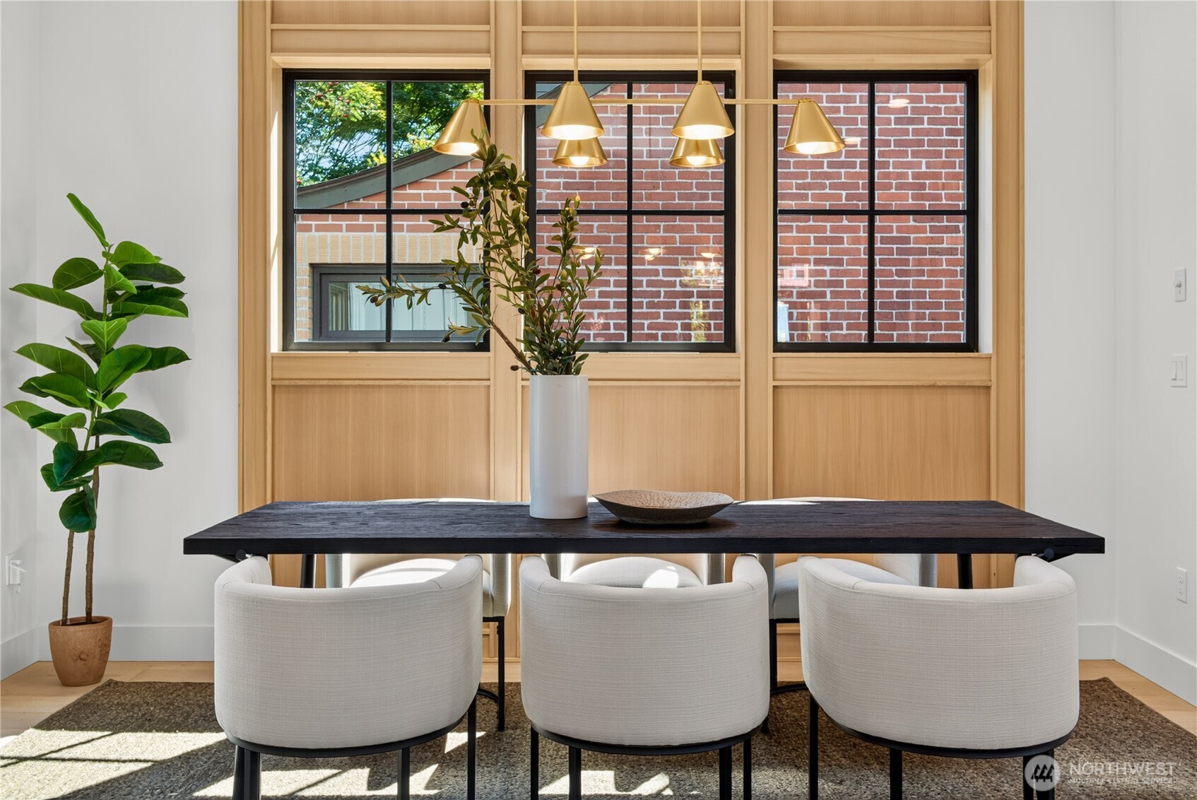 Dining room with a dark table, upholstered chairs, pendant lights, and tall windows framed in light wood.