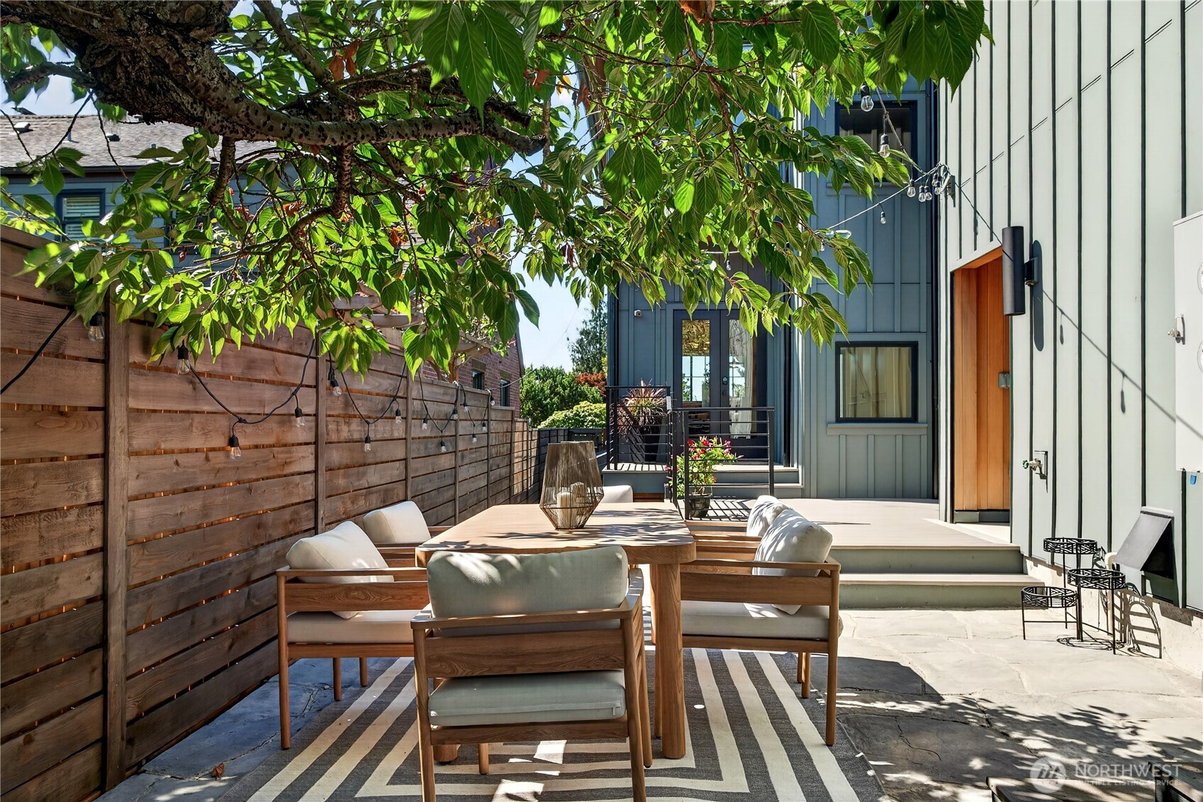 Outdoor dining patio with cushioned wooden chairs beneath a leafy tree beside a modern home.