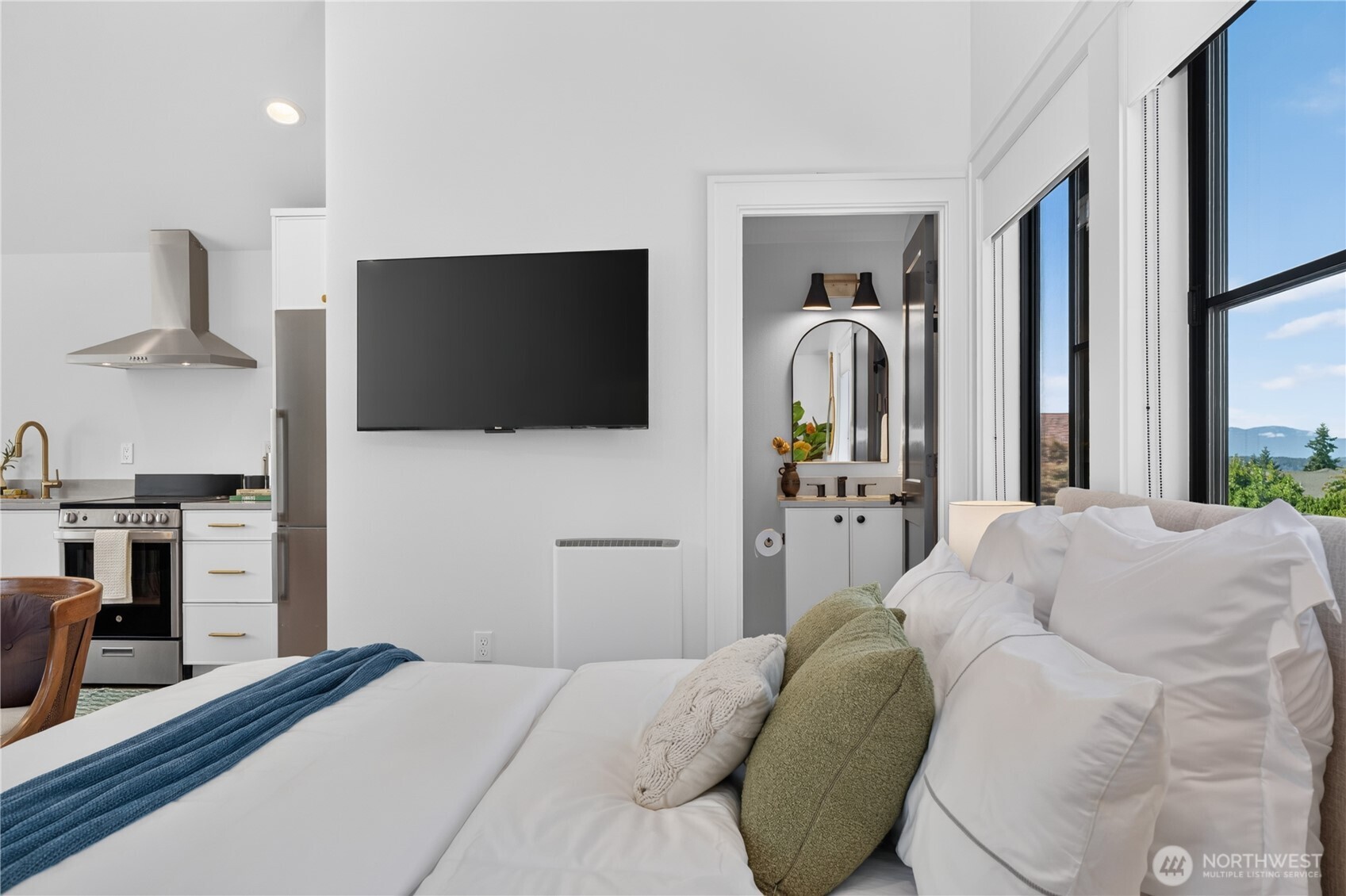 Bright bedroom with a kitchenette, wall-mounted TV, and large windows overlooking distant hills.